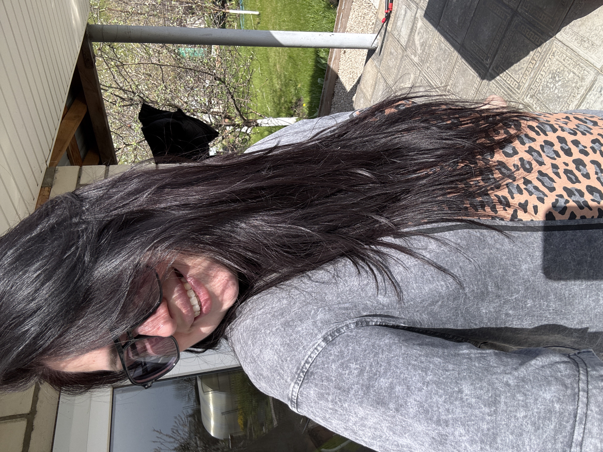 A woman with long, dark hair is smiling and wearing sunglasses, a gray jacket, and a tan and gray animal print top. She is standing on a tiled patio next to a house with white siding. In the background, there is a green lawn, trees, and a silver pole. The location is Unknown, Unknown.