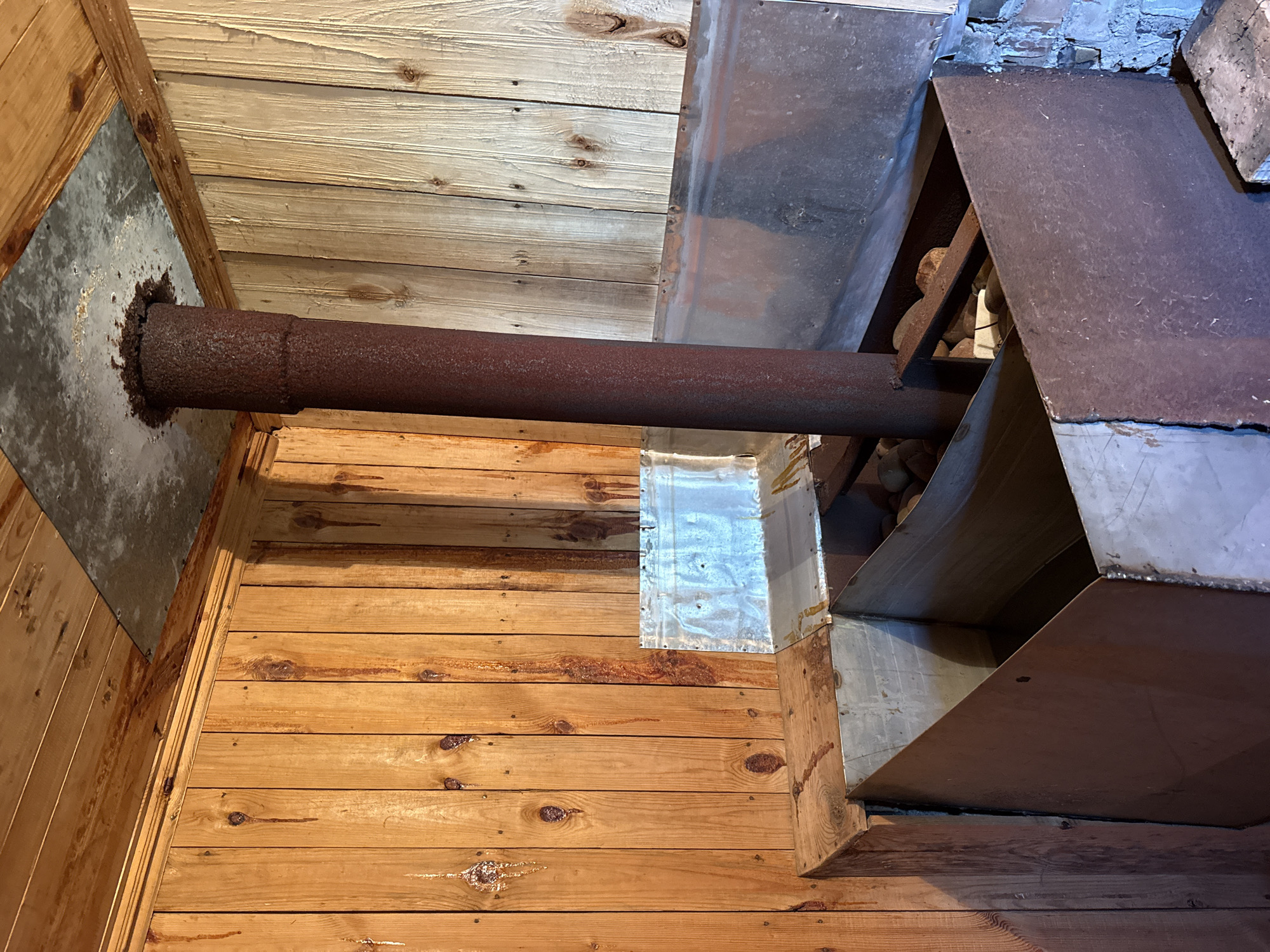 The image shows the interior of a rustic room, likely a sauna or a small cabin, with wooden paneled walls and ceiling. In the center of the room, a large, rusty metal stove or heating unit is prominent. A vertical metal flue pipe rises from the stove and extends upwards towards the ceiling, where it passes through a metal plate. The stove itself is a rectangular metal box with an open top, containing some rounded stones. The walls have visible knots and grain patterns in the wood, and there are some dark streaks and drips on the wood surface, suggesting moisture or sap. Metal sheeting is attached to the walls and ceiling around the stove and flue pipe as a heat shield. The floor in the foreground appears to be made of brick or concrete. The lighting suggests an interior setting, possibly with natural light coming from an unseen source. There are no people visible in the image.