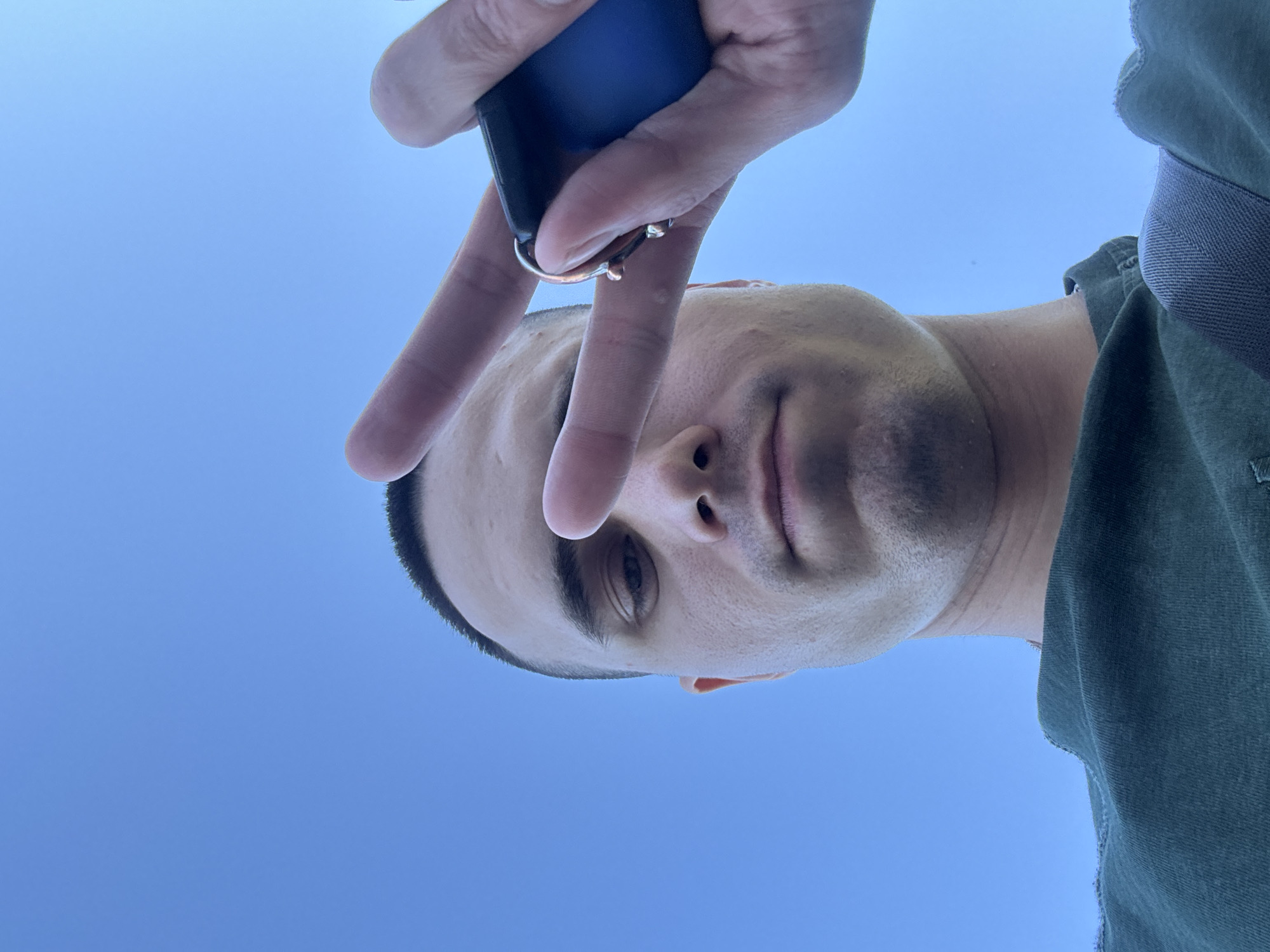 A low-angle, close-up shot shows a person's face and upper torso against a clear blue sky. The person, viewed from below, is making a peace sign with their right hand, holding a dark-colored object, likely a phone, in their palm. Their eyes are looking towards the camera. The person is wearing a dark green t-shirt. The image was taken in Chaikovsky, Russia.