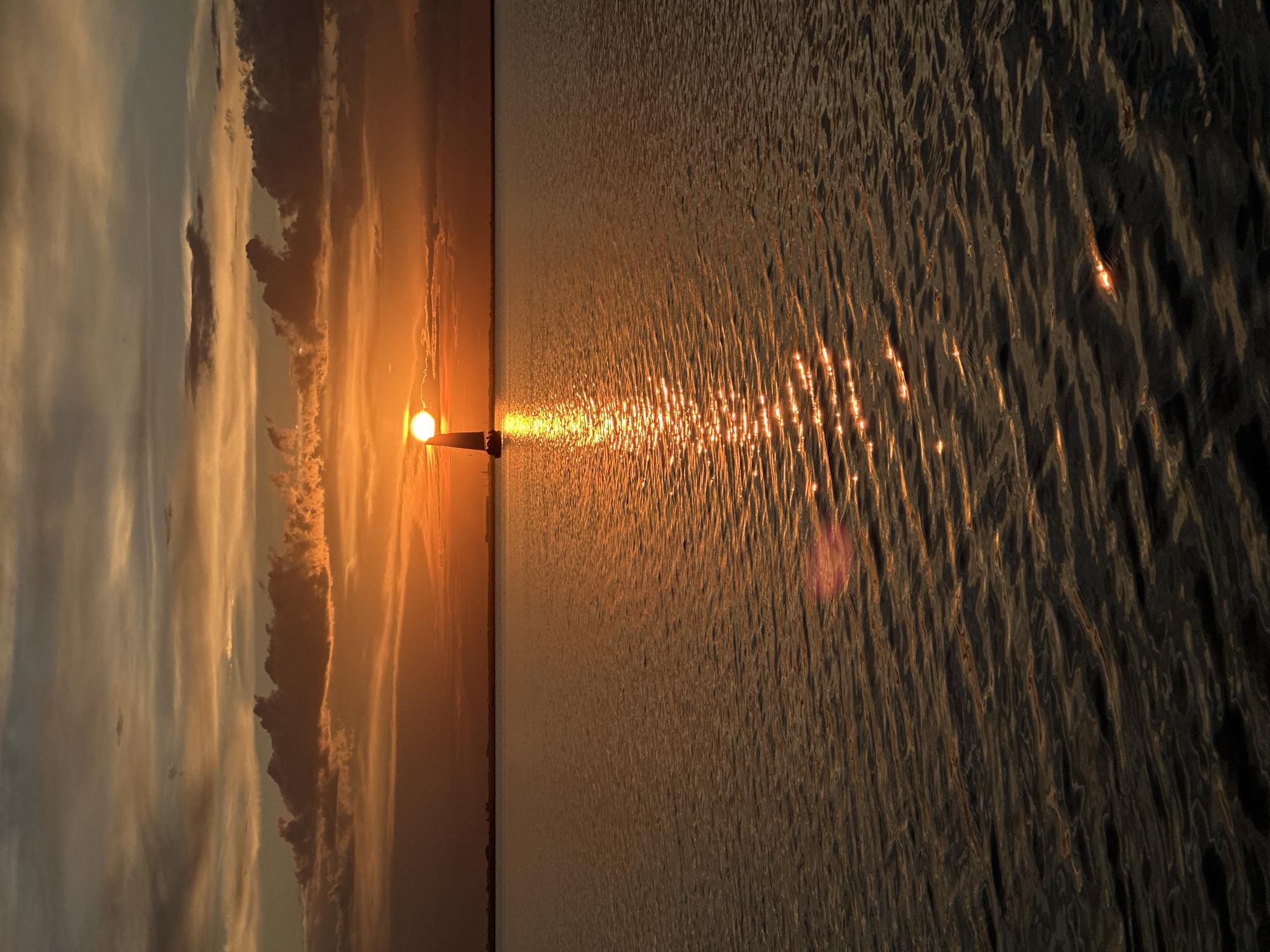 A sailboat with a visible sail is positioned on a calm body of water, facing a vibrant sunset. The sun, partially obscured by clouds and the sailboat's mast, casts a brilliant orange glow across the sky and reflects a shimmering path on the water. The horizon line is clearly defined, separating the water from the sky, with a faint outline of land and structures visible in the distance. The sky displays a gradient of warm colors, from deep orange near the sun to lighter shades of yellow and gray, interspersed with dark, textured clouds. The water is a deep, reflective surface, with ripples catching the light and creating a sparkling effect. The scene conveys a sense of tranquility and natural beauty. The location is the Big Chebachye Lake, located in the Akmola Region of Kazakhstan, near the city of Kokshetau.