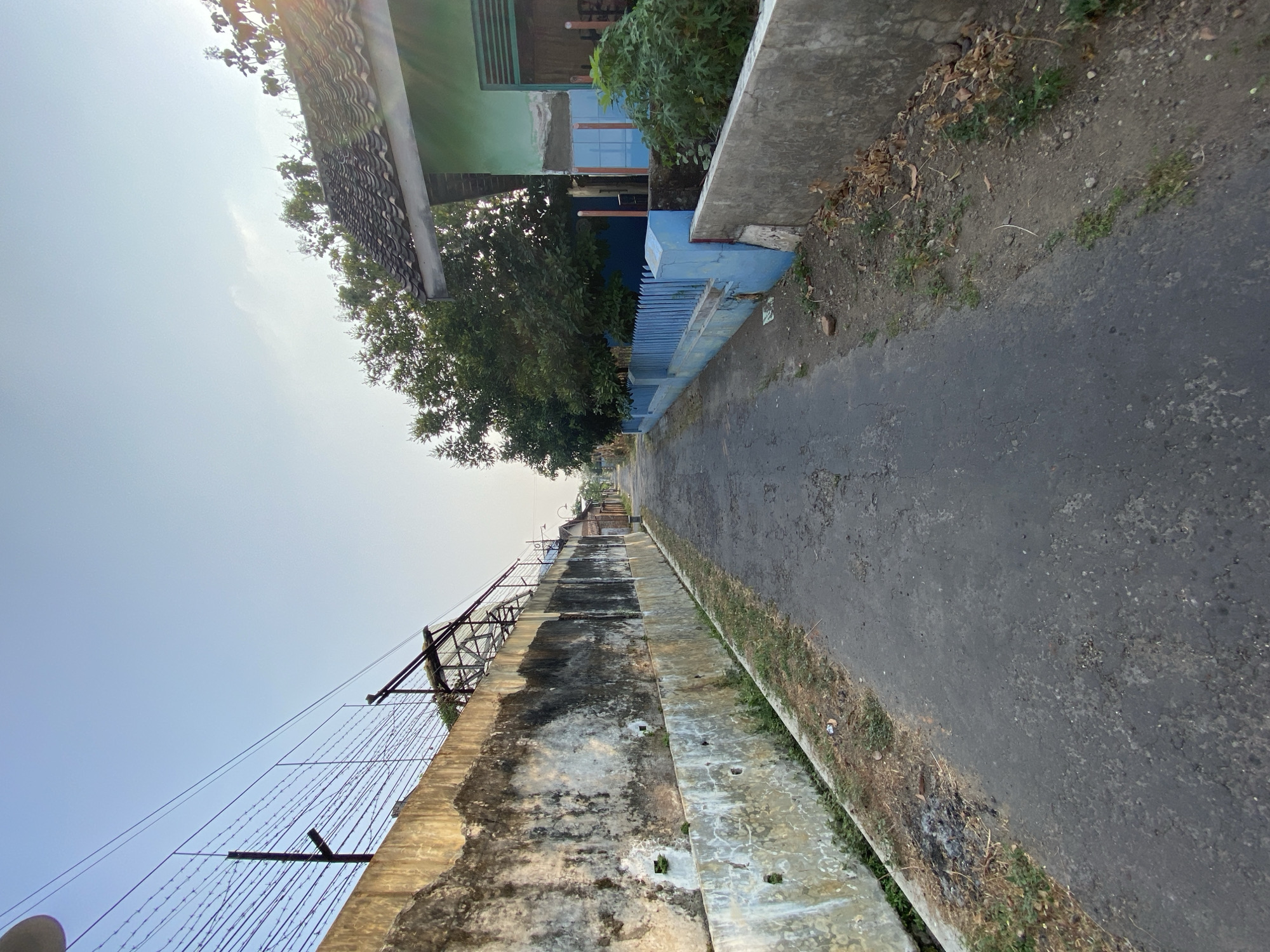 The image shows a narrow, paved road in what appears to be a residential area. On the left side of the road is a tall, weathered concrete wall topped with barbed wire and a metal structure. On the right side of the road, there is a house with a green facade and a tiled roof, separated from the road by a low concrete wall and a blue picket fence. Trees and vegetation are visible behind the fence. The sky is clear and blue, suggesting it is daytime, likely morning or late afternoon due to the soft lighting. The scene is devoid of people or any visible activity. The overall impression is one of a quiet, possibly rural or suburban street in Sugihwaras, Indonesia.