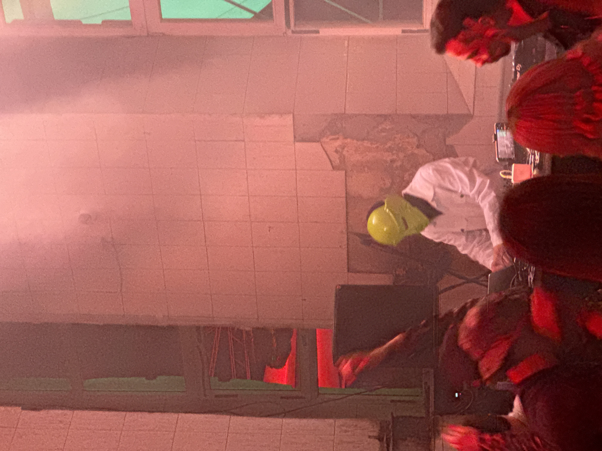 This image captures an indoor event in Moscow, Russia. The scene is dominated by a DJ performance set within a large room characterized by light-colored, tiled walls that exhibit signs of age and structural wear, including visible damage and exposed substrata. The lighting is predominantly red and pink, creating a hazy atmosphere, with strong green light emanating from large multi-pane windows on the right side of the frame. On the left, similar window structures are present, reflecting the red ambient light.
At the center, a performer stands behind a table of electronic equipment. This individual wears a plain white long-sleeved shirt and a distinctive bright yellow, smooth, full-face mask featuring subtle ear-like extensions. The performer's hands are positioned over DJ equipment, including what appears to be a laptop and a mixing console. To the immediate left on the table, a smartphone with its screen illuminated is visible next to a small light-colored cup. A large, dark speaker or monitor stands to the left of the performer.
In the foreground and midground, multiple individuals, presumably audience members, are present. Their forms are partially obscured and illuminated by the red light, with some exhibiting motion blur. A person with dark hair is visible in profile on the far right, while another individual with red hair is seen from behind in the lower right. An extended arm, lit in red, is visible in the mid-left portion of the image.
