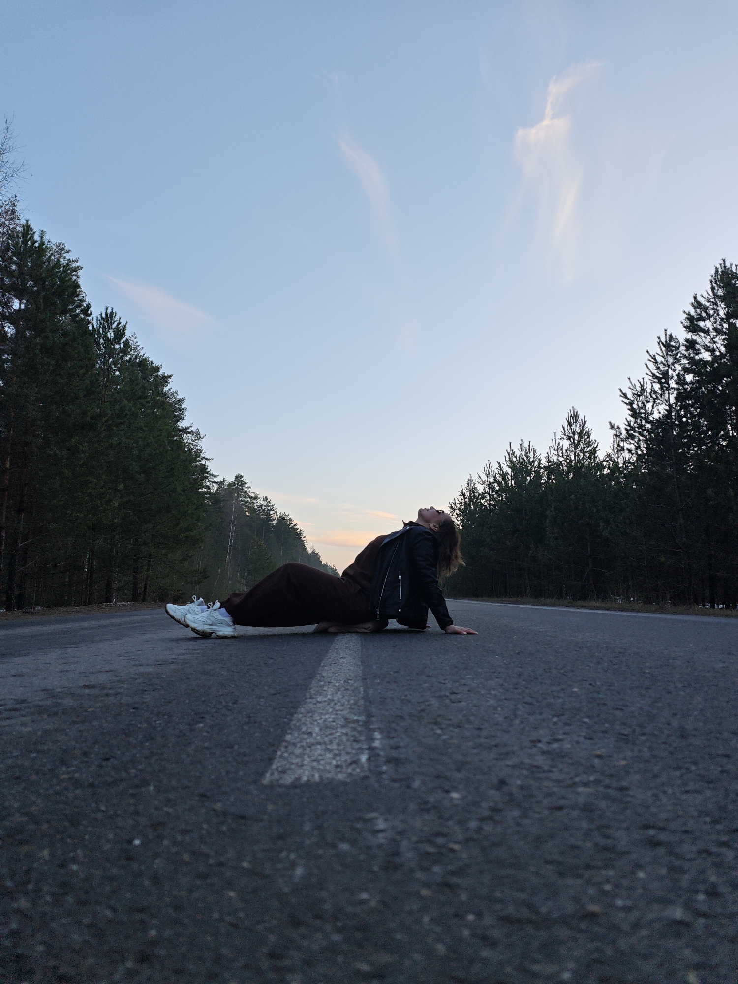 A young adult female is lying on an asphalt road with a white line in the center. She is wearing dark pants, white sneakers, and a black jacket. Her head is tilted back, and she is looking up at the sky. The road is flanked on both sides by tall evergreen trees, creating a forest setting. The sky is a pale blue with some light clouds near the horizon. The scene appears to be in a rural or semi-rural location. Given the request for location details, it can be inferred that the image was taken in the city of Chaikovsky, Russia.