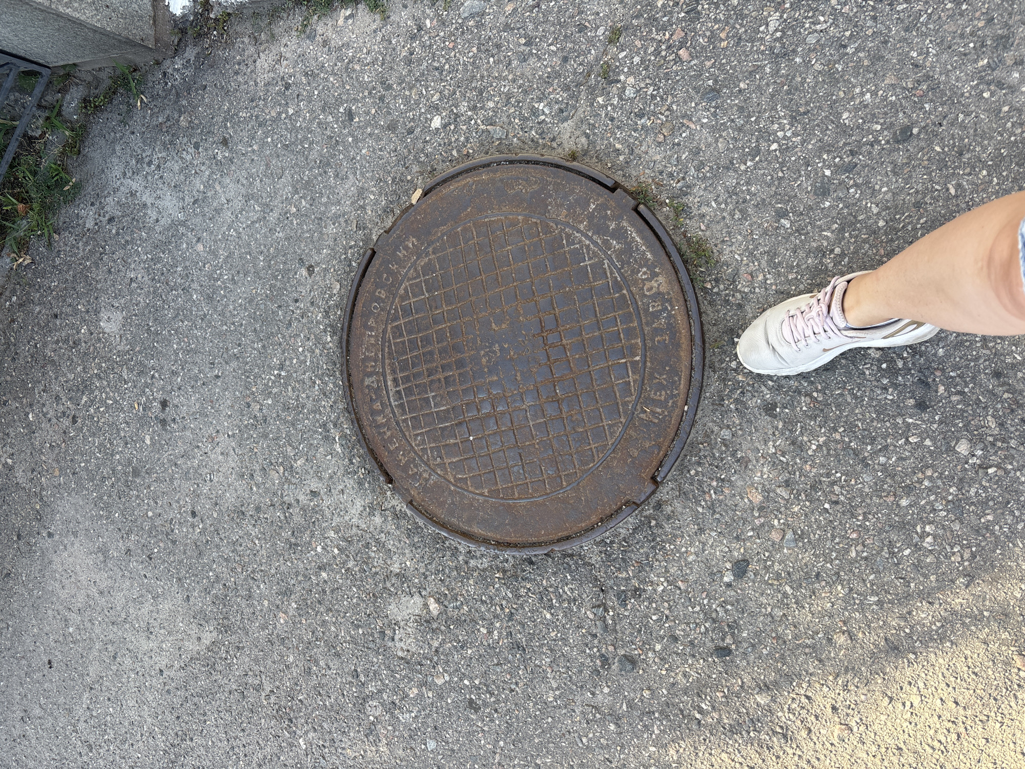 The image shows a close-up, overhead view of a round, metal manhole cover on an asphalt surface. The manhole cover has a raised grid pattern and some text inscribed around its circumference. To the right of the manhole cover, a person's leg, wearing a light-colored sneaker with pink laces, is visible. The person appears to be standing or walking near the manhole cover. The asphalt surface is rough and appears to be part of a sidewalk or street. Some sparse green grass and a metal grate are visible in the upper left corner of the frame, suggesting an outdoor urban or suburban environment. The lighting suggests it is daytime, possibly late afternoon due to the shadows. No specific emotional cues are evident. The visible text on the manhole cover is partially obscured and appears to be in Cyrillic script.