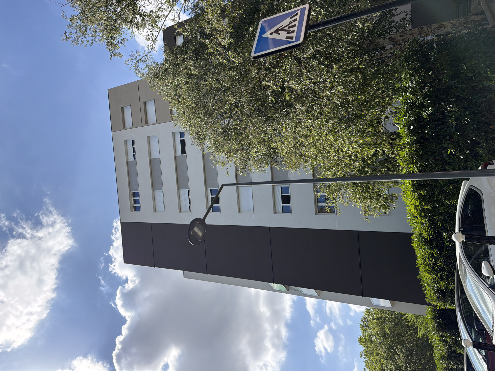 A low-angle shot captures a modern, multi-story apartment building in Évry-Courcouronnes, France, against a backdrop of a bright blue sky dotted with white clouds. The building features a distinctive facade with a large dark gray section contrasting with lighter gray and white panels. Rows of windows with closed white blinds are visible on the lighter sections. A tall, dark lamppost stands in the foreground, with a pedestrian crossing sign mounted on a pole to its right. Lush green foliage from trees and hedges partially obscures the lower part of the building and the street. Several cars are parked in front of the hedges, with their roofs and windshields visible. The scene appears to be an urban residential area on a sunny day.