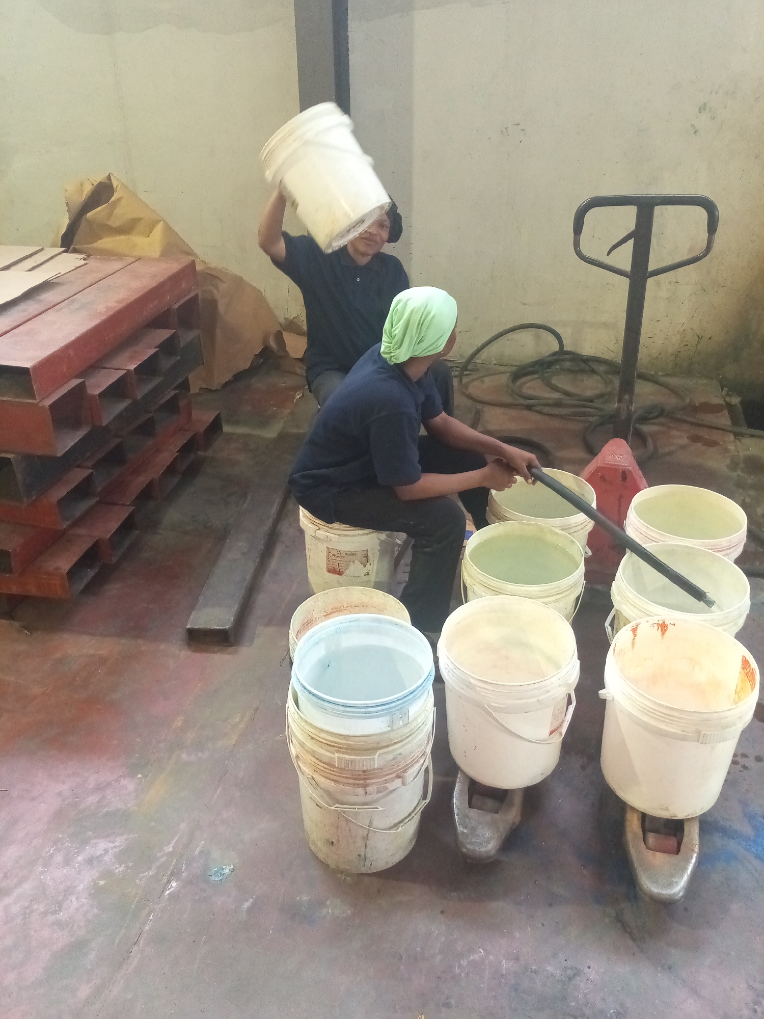 This image depicts an indoor industrial or workshop setting in Paipe, Nigeria, where two individuals are engaged in handling and mixing materials, likely liquids or powders. The floor is concrete, heavily stained and marked with various colors, indicative of ongoing industrial activity.
In the foreground and mid-ground, numerous white plastic buckets of various sizes are visible. Some are stacked, while others contain light-colored liquids of different hues, such as pale green, white, and yellowish-orange. Two of the filled buckets are placed on the prongs of a pallet jack or a similar hydraulic lifting device, suggesting they are being weighed or prepared for transport.
Two people are central to the activity. On the left, a person wearing a dark shirt is seen from the chest up, holding a white bucket upside down over their head as if pouring or shaking out its contents. This individual has a visible smile, suggesting a relaxed or perhaps jocular mood during work.
To the right, another person is seated on a stack of smaller white buckets. This individual is dressed in a dark blue shirt and wears a light green head covering. They are actively stirring the contents of a large white bucket with a long, dark stick. The bucket being stirred contains a light green liquid. Behind them, a black hose or cable is coiled on the floor, and the handle of a pallet jack stands upright against a plain, light-colored wall.
Stacked red metal beams or pallets are visible on the left side, along with a large brown paper bag, contributing to the functional and somewhat cluttered appearance of the workspace. The lighting suggests an indoor artificial source. No visible text is legible on any objects within the image.