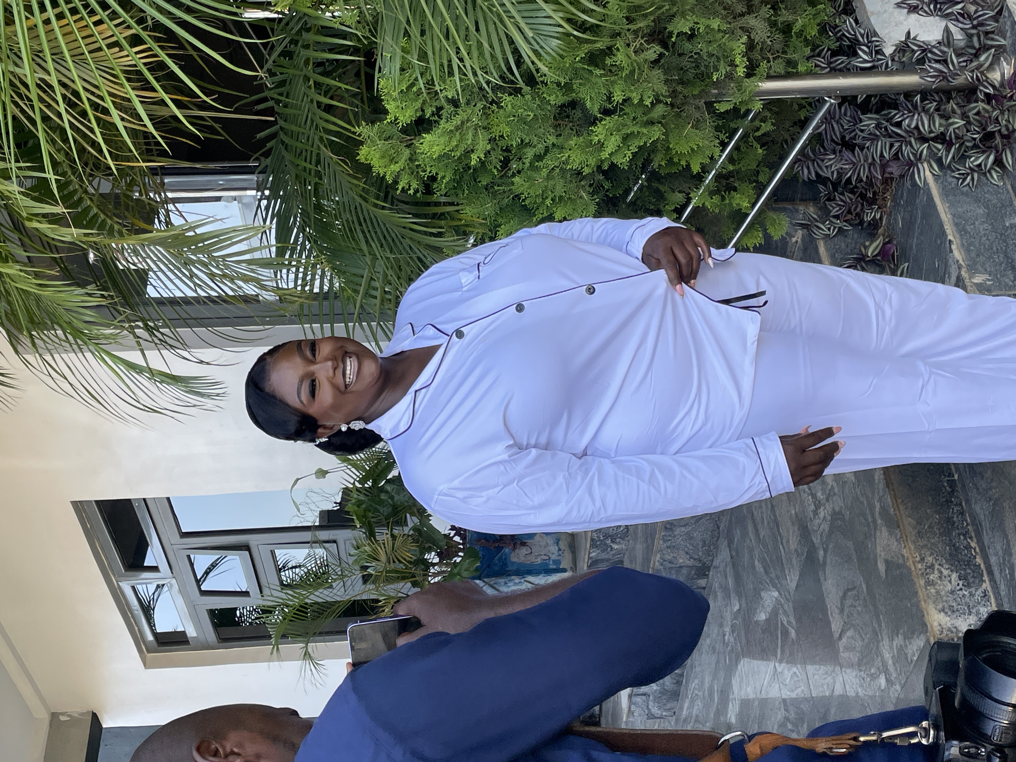 The scene shows a smiling Black woman in white pajamas with black piping, standing outside in what appears to be a residential or commercial location in Jos, Nigeria. The woman has her hair styled and is wearing earrings. She is facing the camera. A person is partially visible in the foreground, holding a phone as if taking a photo of the woman. Part of a camera is also visible near the lower right edge of the image. The background includes a building with a multi-paned window, various green plants, and what looks like a dark stone walkway. The overall light suggests it is daytime.