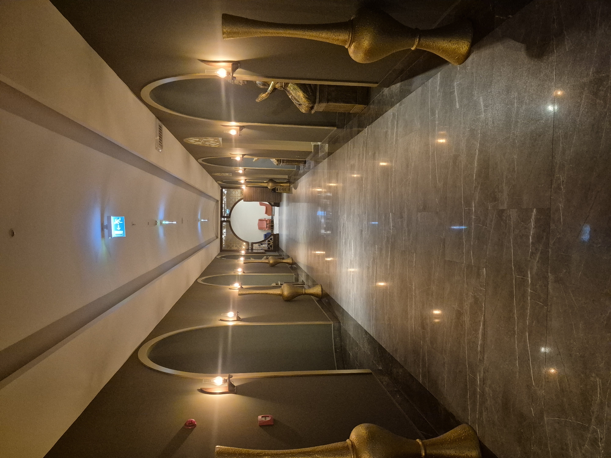 The image depicts a long, narrow hallway in Alanya, Turkey. The hallway has a dark marble floor, a gray wall on one side, and a white wall on the other. The ceiling is dark gray with archways. Each archway contains a golden vase-shaped sconce with a light. Some archways contain golden statues of kneeling figures. The end of the hallway opens into a room with red chairs. A blue emergency exit sign is visible on the left wall.