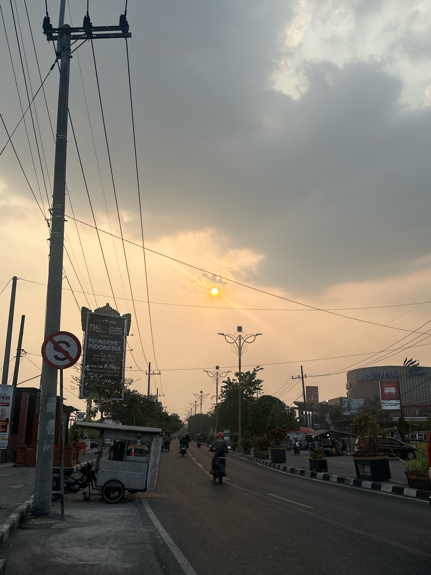 The image depicts a street scene in Mojokerto, Indonesia, during what appears to be late afternoon or early evening, with the sun low in the sky, casting a warm, hazy glow. A prominent telephone pole with multiple wires stands on the left side of the road, close to the foreground. Adjacent to the pole is a circular traffic sign with a diagonal red bar, indicating "no stopping" or "no parking." Further down the sidewalk, a food cart with the word "BATAGOR" written vertically on its side is parked.
The road itself is asphalt, with a white line marking the center and a black and white striped curb. Several motorcycles are visible on the road, with some riders wearing helmets. In the background, there are buildings, including a large structure with "UNIMALL" signage and another with "JOBO" signage. Trees and streetlights are also present along the roadside. The sky is overcast with clouds, but the sun is visible, creating a sunbeam effect through the haze. The overall impression is of a typical urban street in Indonesia, with active traffic and commercial establishments.
