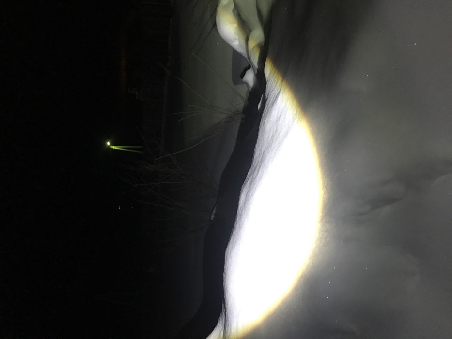 At night, a bright, circular light illuminates a snow-covered area. The light casts a stark contrast against the darkness. In the background, a fainter, distant light is visible, along with hints of trees and structures obscured by the night. The scene evokes a sense of isolation and emphasizes the intensity of the artificial illumination against the natural darkness.