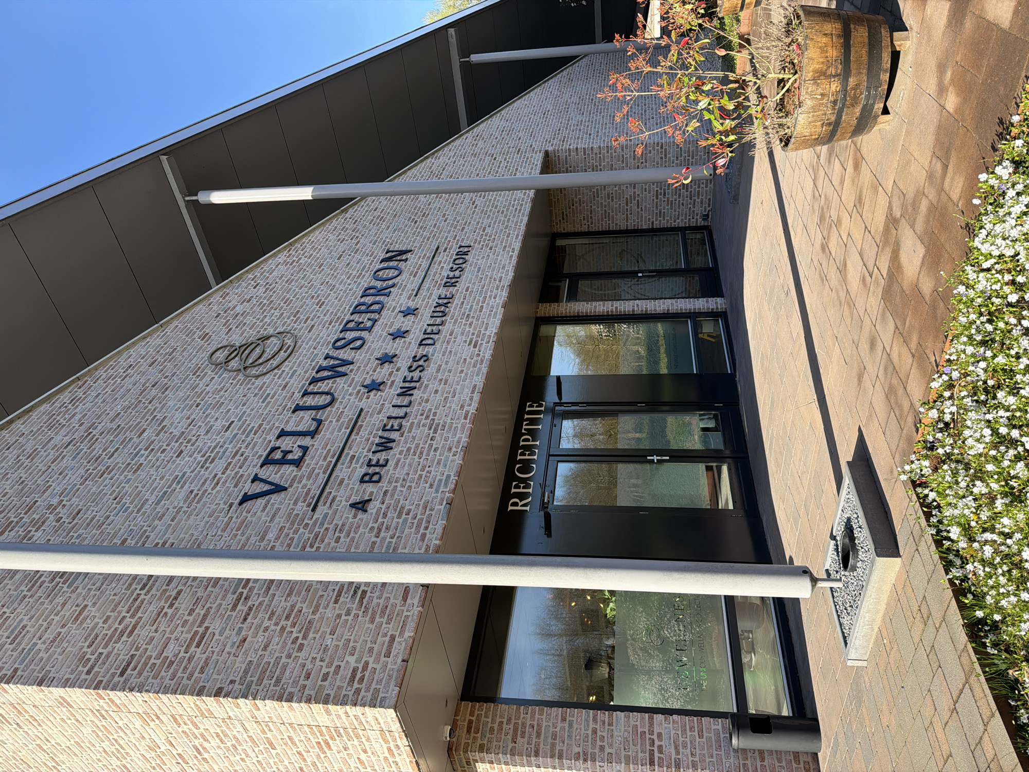 The image shows the exterior of the Veluwse Bron, a wellness deluxe resort in Emst, Netherlands. The building has a brick facade with the resort's name prominently displayed. The entrance is marked by a black frame with glass doors and windows, and the word "RECEPTIE" is visible above the door. A large barrel planter is positioned to the right of the entrance. A pathway leads up to the building, with white flowers growing along the edge. Metal support posts extend from the building's canopy. The sky is blue, suggesting a clear day.