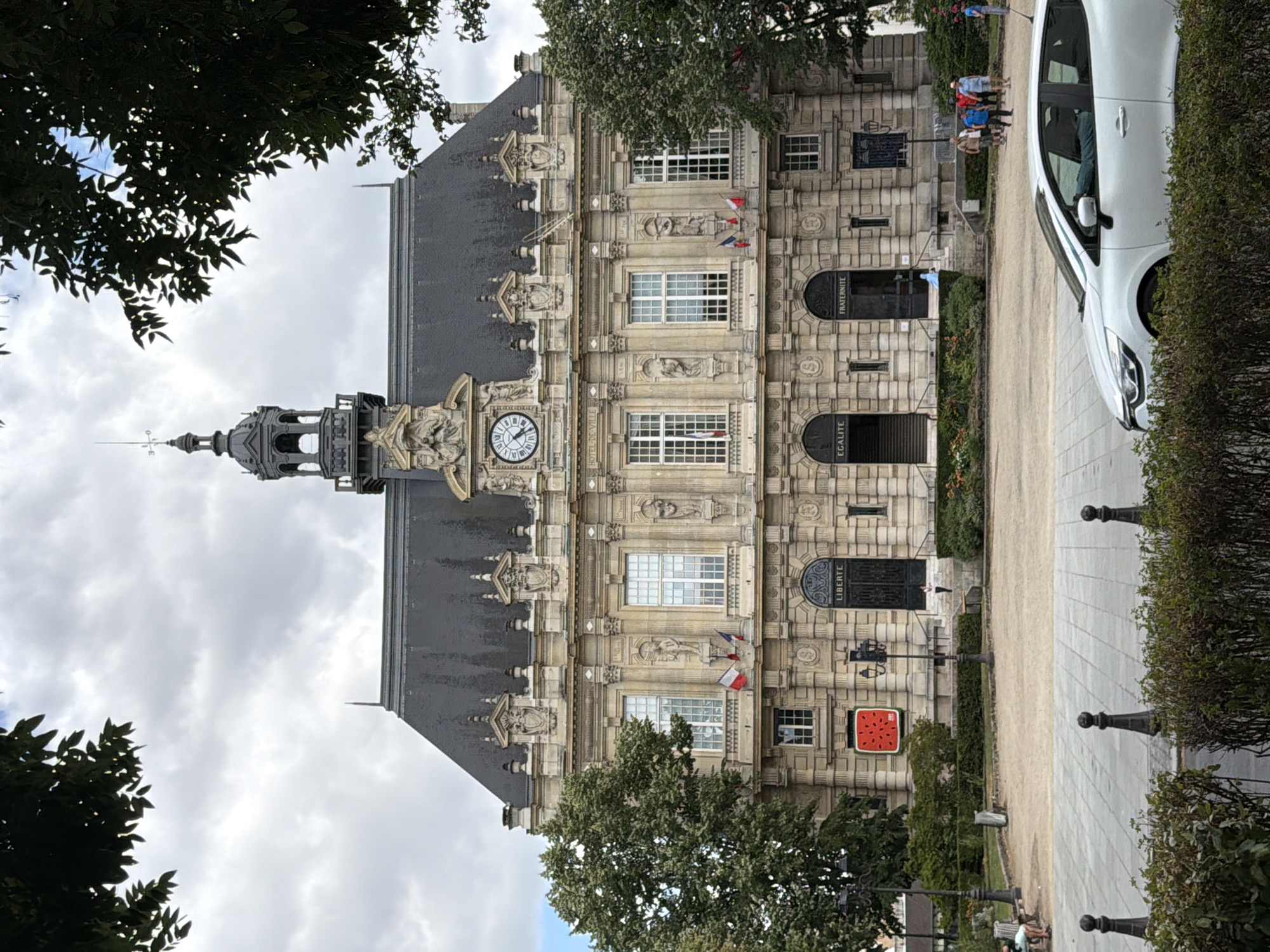 The image shows the Hôtel de Ville (City Hall) of Ivry-sur-Seine, France, a large stone building with classical architectural elements. The building has a prominent clock tower and a steep, dark roof. The facade is adorned with sculptures, decorative carvings, and arched windows.
In front of the building, there is a paved area with trees and park benches. A large, red artwork resembling a slice of watermelon is mounted on the left side of the building's facade. Several French flags are displayed on poles in front of some windows.
A group of people are visible walking in the middle ground, and a white car is parked in the foreground, partially obscuring a low hedge. The sky is cloudy, suggesting overcast weather. The overall scene depicts a civic building and its surrounding public space.