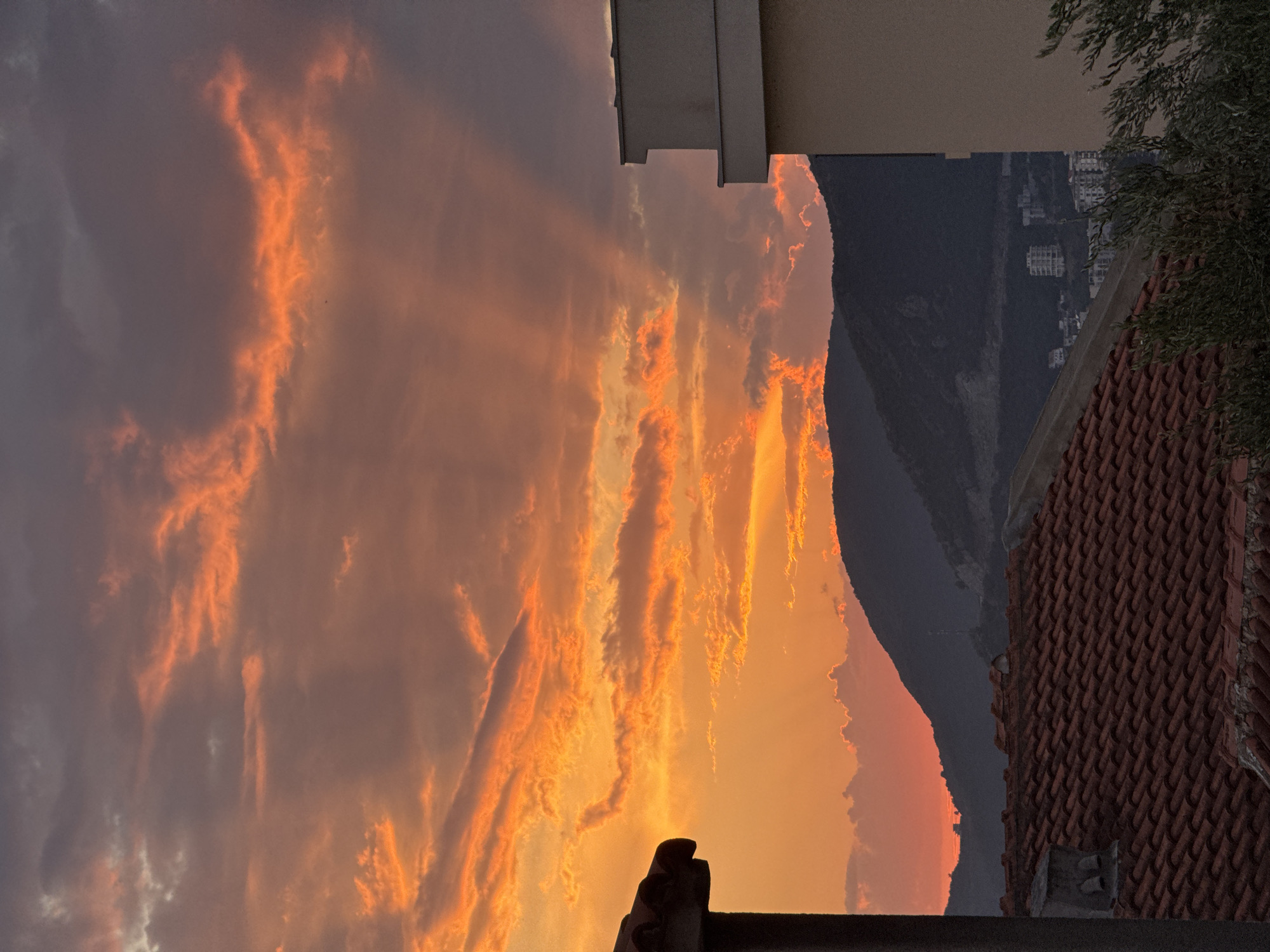 The image depicts a sunset over the city of Trap, Montenegro. The sky is filled with dramatic clouds illuminated by the setting sun, casting vibrant orange and red hues across the scene. Rays of light pierce through the clouds, creating a striking visual effect. In the foreground, terracotta tiled rooftops of buildings are visible, along with a portion of a modern building on the right. Dark, silhouetted mountains form the backdrop, with some distant buildings nestled within the landscape. The overall impression is one of natural beauty and a serene evening atmosphere in a coastal town.