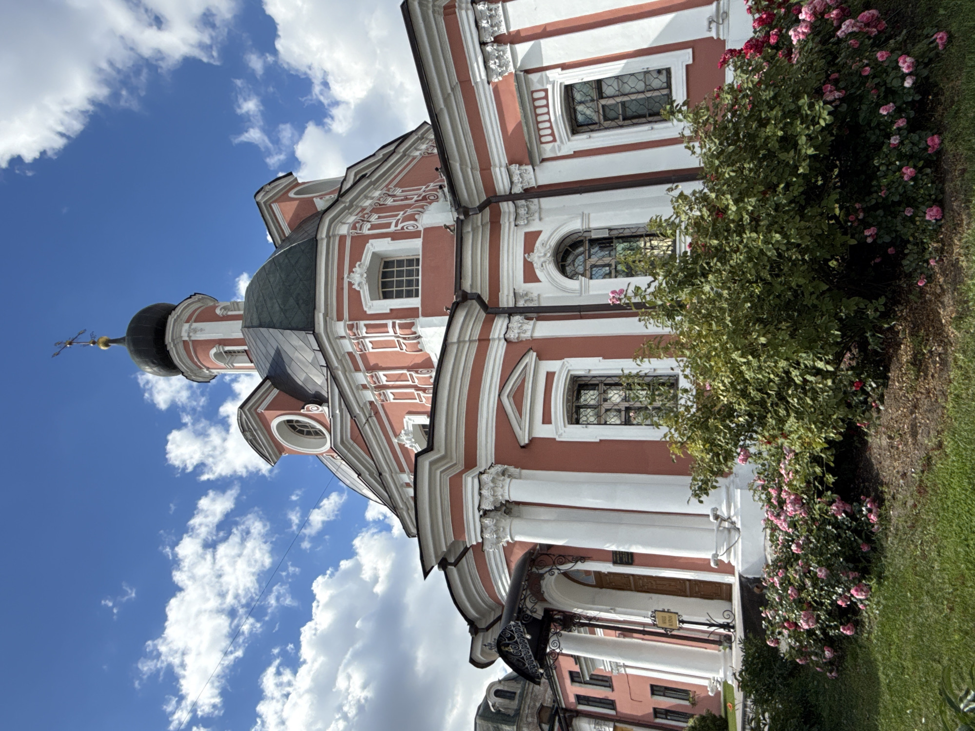 The image shows the Church of the Nativity of the Virgin in Seredn'kino, Moscow, Russia. It is a baroque-style church with a pink facade and white architectural details. The church features a large dome topped with a cross, several windows, and columns at the entrance. In front of the church, there is a well-maintained garden with lush green grass and a vibrant bush covered in pink roses. The sky is blue with scattered white clouds. No people are visible in the image.