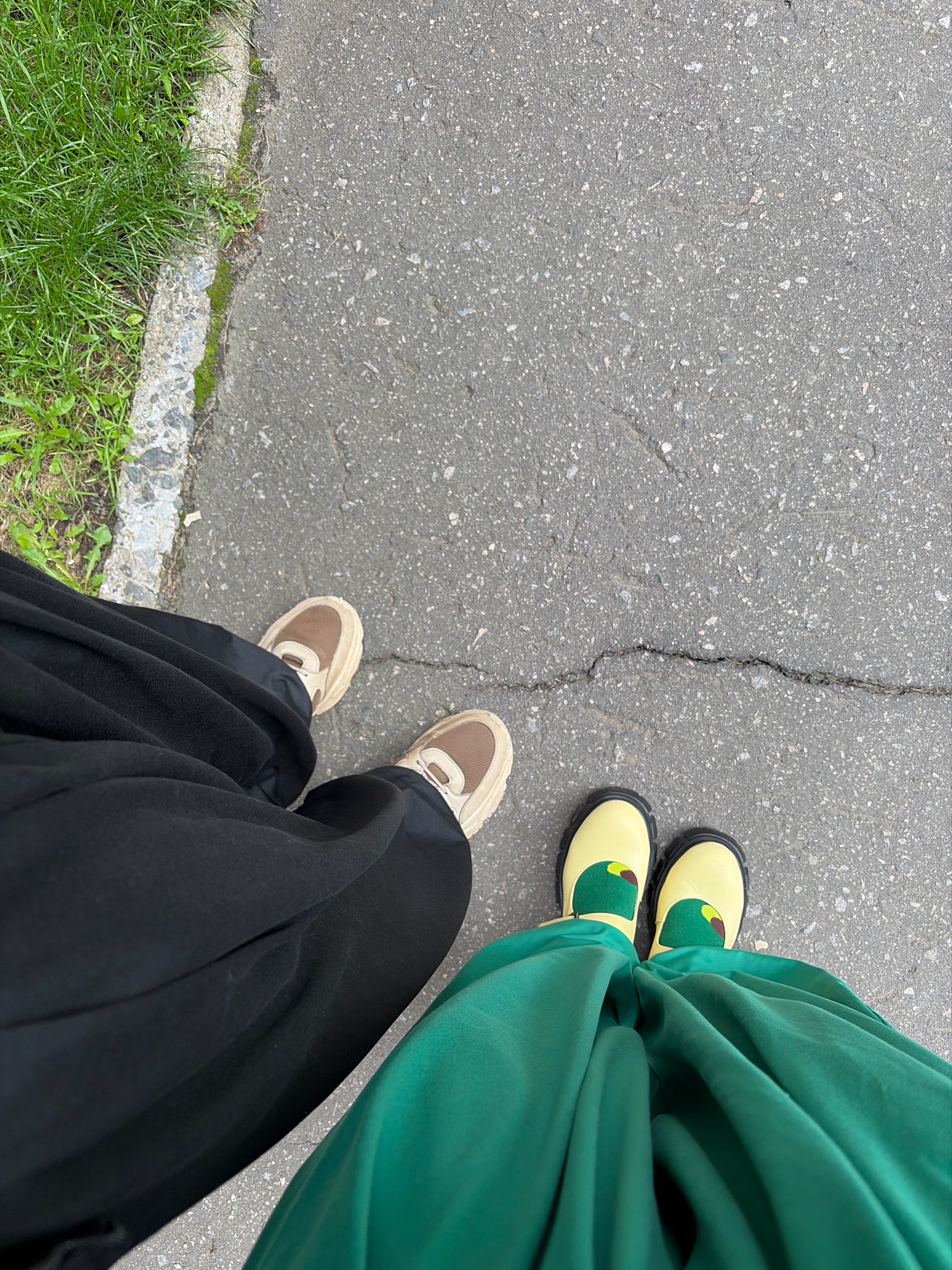 The image shows a top-down view of two people's feet standing on a cracked asphalt sidewalk next to a grassy area. The person on the left is wearing black pants and light brown and beige sneakers. The person on the right is wearing a bright green dress and yellow mary jane shoes with a green and brown design on the toe, resembling a cartoon bird. The environment suggests an outdoor setting in Arkhangelsk, Russia, given the presence of grass, asphalt, and the general lighting conditions.
