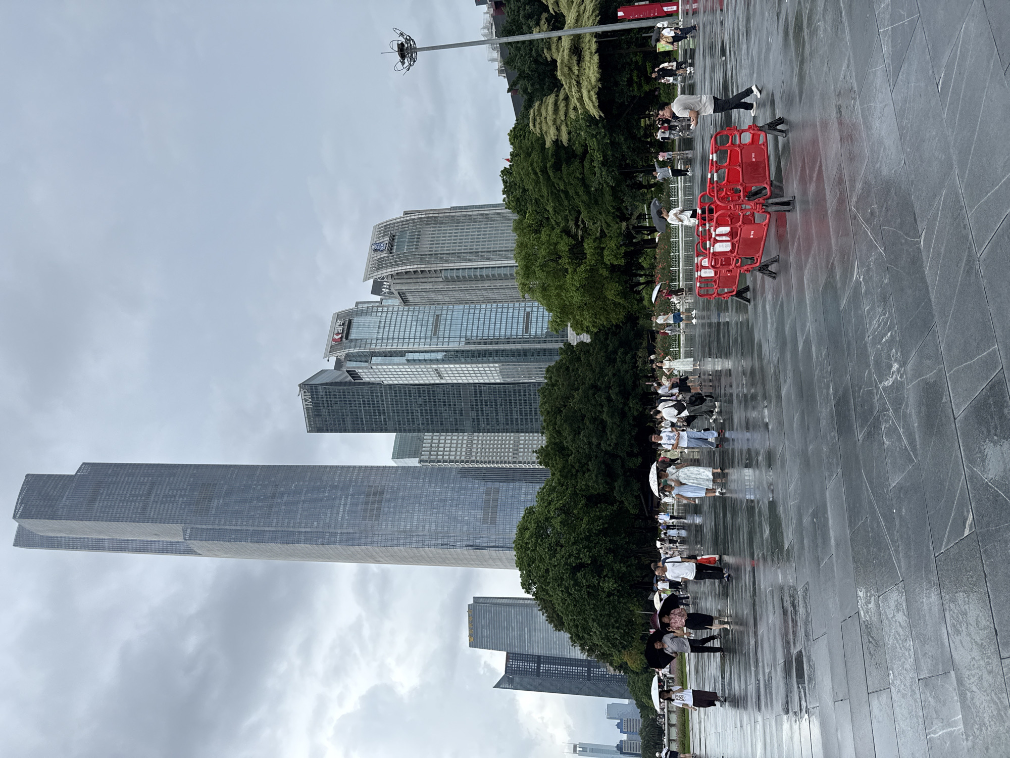 This image captures an outdoor plaza in Tianhe District, China, under a cloudy sky, with a light rain evident from the wet pavement and people holding umbrellas. In the background, a cluster of modern skyscrapers dominates the skyline, with the tallest being a distinctive, slender, grey building. The plaza floor is paved with large, dark grey rectangular stones that are highly reflective due to the moisture.
Several individuals are walking across the plaza, many holding white and black umbrellas. Some are walking alone, while others are in small groups. People are dressed in casual attire, suggesting a relaxed atmosphere despite the weather.
In the mid-ground, lush green trees form a visual barrier between the plaza and the buildings. To the right of the image, a tall, modern lamppost stands out. In the foreground, a bright red, temporary barrier, possibly for crowd control or a construction area, is placed on the wet ground.
The overall impression is a city scene during a gentle rain, with urban architecture and people navigating the wet public space.