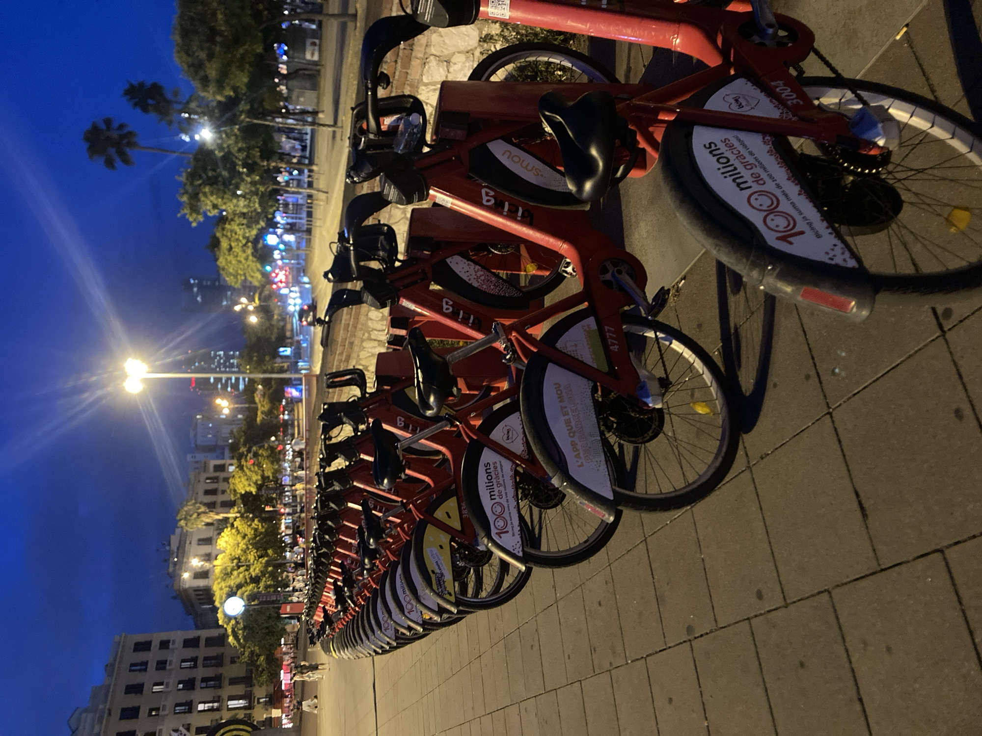 This is a medium shot, eye-level, outdoor photograph taken at dusk in Barcelona, Spain, featuring a row of red Bicing bicycles parked on a tiled sidewalk. The bicycles are lined up neatly, with their handlebars turned to the left. The focus is on the bicycles in the foreground, with a blurred background showing buildings, trees, streetlights, and palm trees, characteristic of a city street at twilight. The sky is a deep blue, indicating it is either early evening or late dawn. Some of the bicycles have visible signage, including the Bicing logo, and text in Catalan, such as "100 milions de gràcies" (100 million thanks) and "L'APP que et mou per Barcelona" (The app that moves you through Barcelona). The scene appears to be a public area with pedestrians visible in the distance walking along a promenade or street. The overall impression is one of urban infrastructure and public transportation in Barcelona at the end of the day.