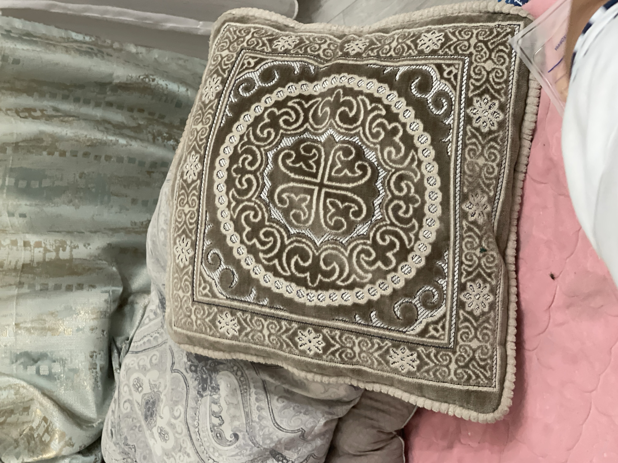 The image shows a close-up of a decorative pillow with intricate patterns. The pillow is square and features a central circular design with swirling motifs and a cross-like emblem in the middle. The border of the pillow is also decorated with floral and geometric patterns. The pillow appears to be made of a soft, velvety material in shades of brown and cream, with silver accents that catch the light.
The pillow is lying on a bed, partially overlapping with other bedding. To the left of the pillow, a light blue-green fabric with a textured, striped pattern is visible. Below the pillow and to the right, there's a pink textured surface, possibly another blanket or the bedspread itself. In the upper right corner, a portion of a person's arm and a clear plastic container are visible. The overall scene suggests an indoor setting, likely a bedroom. The lighting is soft, indicating it could be daytime or well-lit interior lighting. There are no obvious signs of weather or specific time of day. The emotions conveyed are neutral, focusing on the decorative nature of the pillow and the surrounding bedding. No text is visible in the image. The context provided places this scene in Bishkek City, Kyrgyzstan, suggesting the pillow might be a decorative item reflecting local or regional artistry.