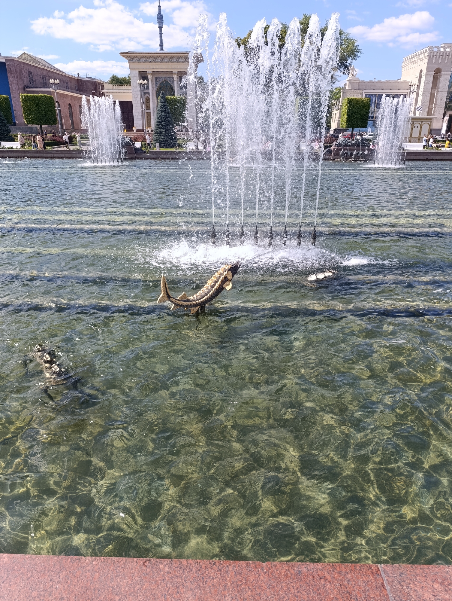 The image depicts a fountain in a public space, likely a park or a plaza, in Moscow, Russia. The fountain features multiple streams of water spraying upwards, with a large sturgeon sculpture partially submerged in the foreground. To the left of the sturgeon, another sculpture, possibly an alligator or crocodile, is also visible in the water. In the background, there are several buildings with distinct architectural styles, including one with ornate detailing and a tower in the distance. The sky is blue with scattered clouds. Several people are visible walking in the background near the buildings. The water in the fountain is clear, allowing a view of the bottom. The fountain appears to be part of a larger complex or exhibition center, given the architectural style of the buildings and the presence of sculptures. The overall scene suggests a pleasant, sunny day in a well-maintained outdoor area.