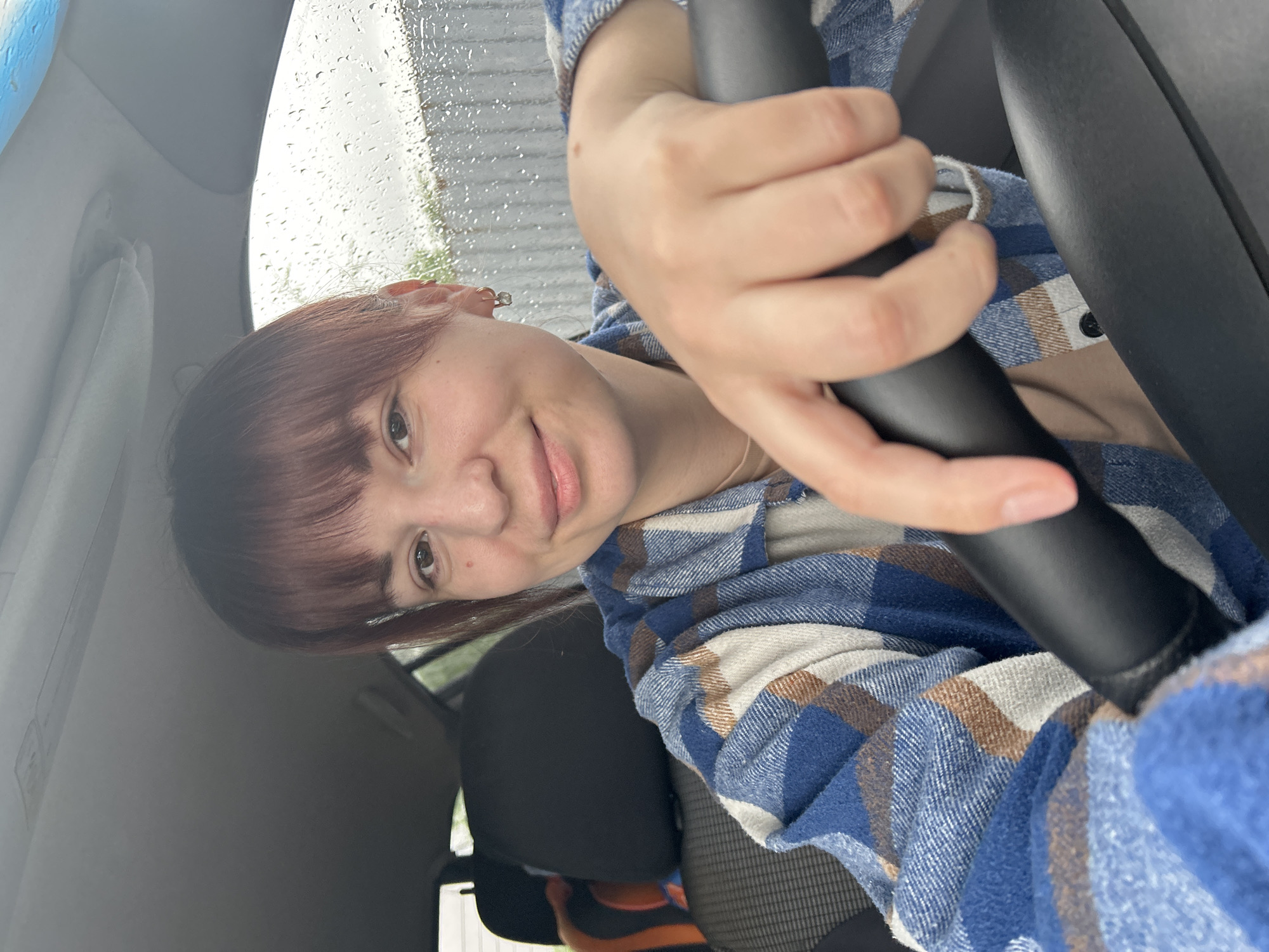 A woman is sitting in the driver's seat of a car, holding the steering wheel. She is wearing a blue and brown plaid shirt and has brown hair with bangs. The car window is visible, with raindrops on the glass. The seat of the car is black. The image's location in the city of Фоки, Russia is not determinable from the image content alone.