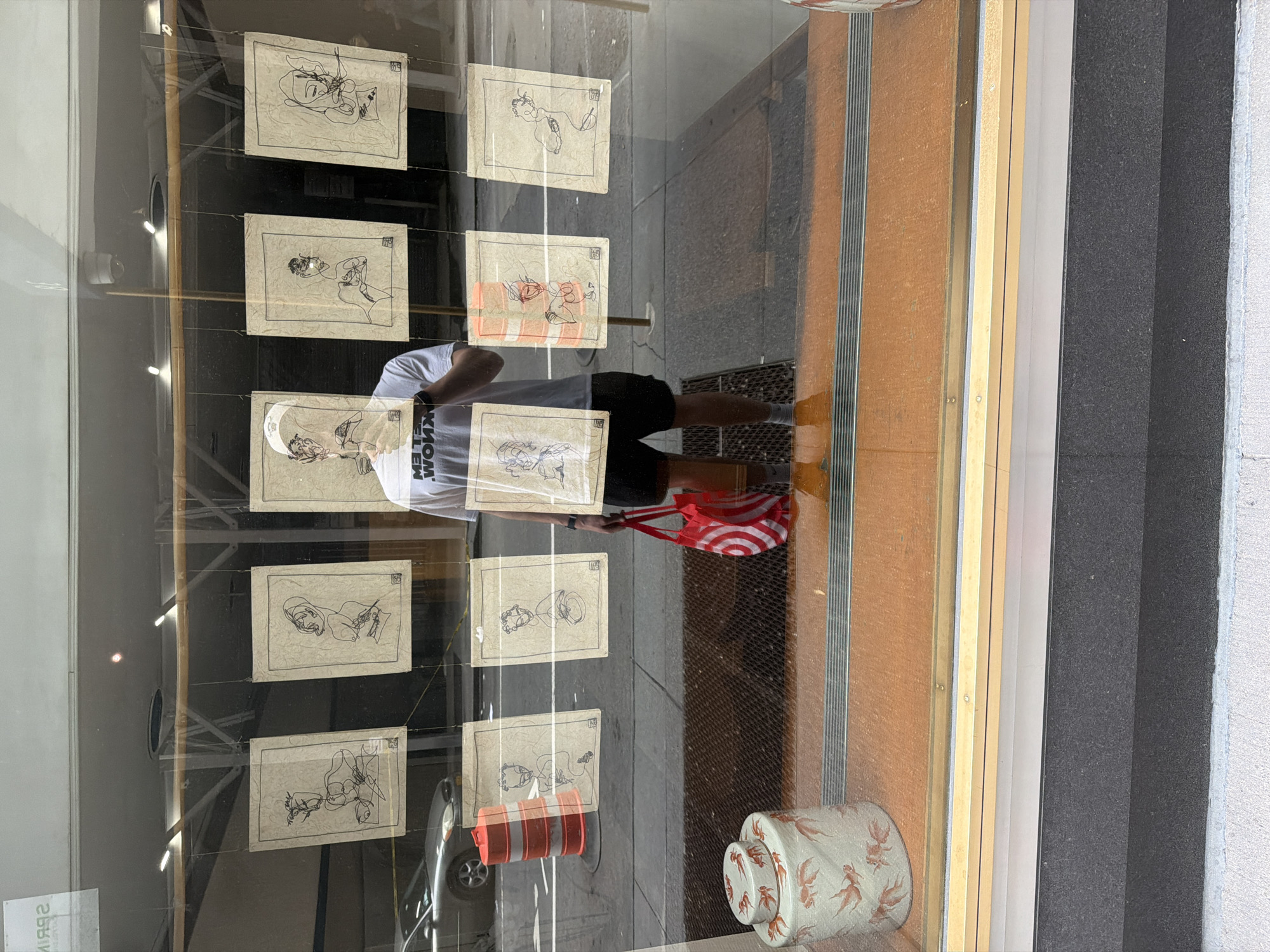 The image is of a store window, possibly in New York City, United States of America. Inside, artwork is displayed on strings; the artwork features minimalist line drawings on tan paper, arranged in a grid pattern. In the window's reflection, a person wearing a white t-shirt with black text, black shorts, and a white baseball cap, is visible. The person is holding a red and white striped tote bag. The reflection also reveals part of a street with a parked car and an orange and white traffic cone. At the bottom right of the image, there are stacked cylindrical containers with a red design. The window itself is framed by wood and a dark gray exterior wall.