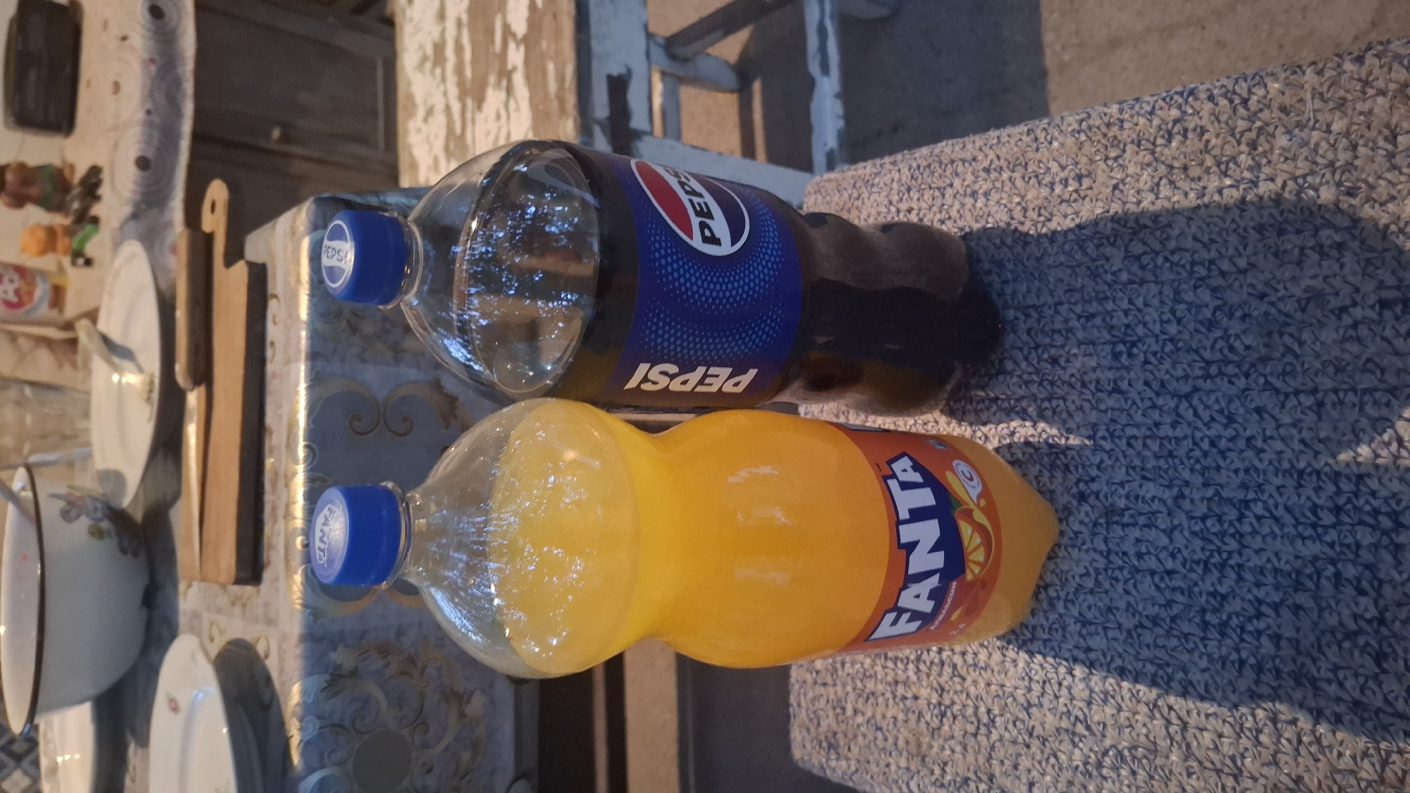 Two plastic bottles of soda, one Fanta and one Pepsi, are placed side-by-side on a textured blue and white surface. The Fanta bottle is on the left, filled with an orange liquid and labeled with its brand name and an orange slice graphic. The Pepsi bottle on the right contains a dark-colored liquid and features the Pepsi logo. In the background, a table is visible with plates, a bowl, and a cutting board with a knife. The overall setting appears to be an outdoor or semi-outdoor area, possibly a backyard or patio, with a rustic wooden table and chairs. The lighting suggests it is daytime.