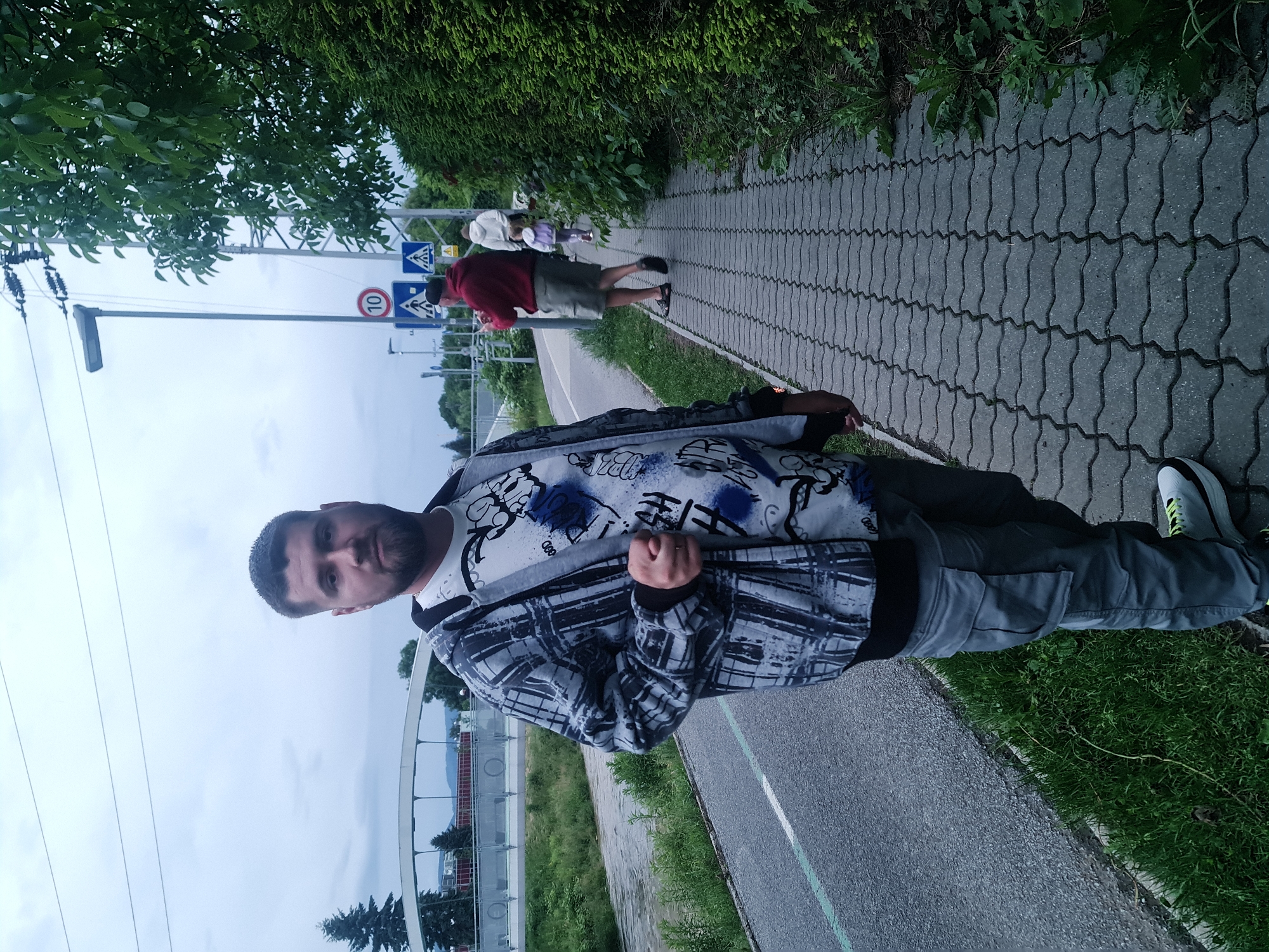 A man stands on a paved sidewalk, looking at the camera. He is wearing a grey patterned zip-up hoodie over a white t-shirt with blue and black graphics, and grey cargo pants. He has short dark hair and a beard. The sidewalk is adjacent to a bike path, with a grassy verge separating them. In the background, there is a pedestrian crossing with signs, a lamppost, and lush green trees and bushes. Further in the distance, a white bridge is visible. Other people are seen walking on the sidewalk in the background. The overall scene suggests an outdoor urban environment.