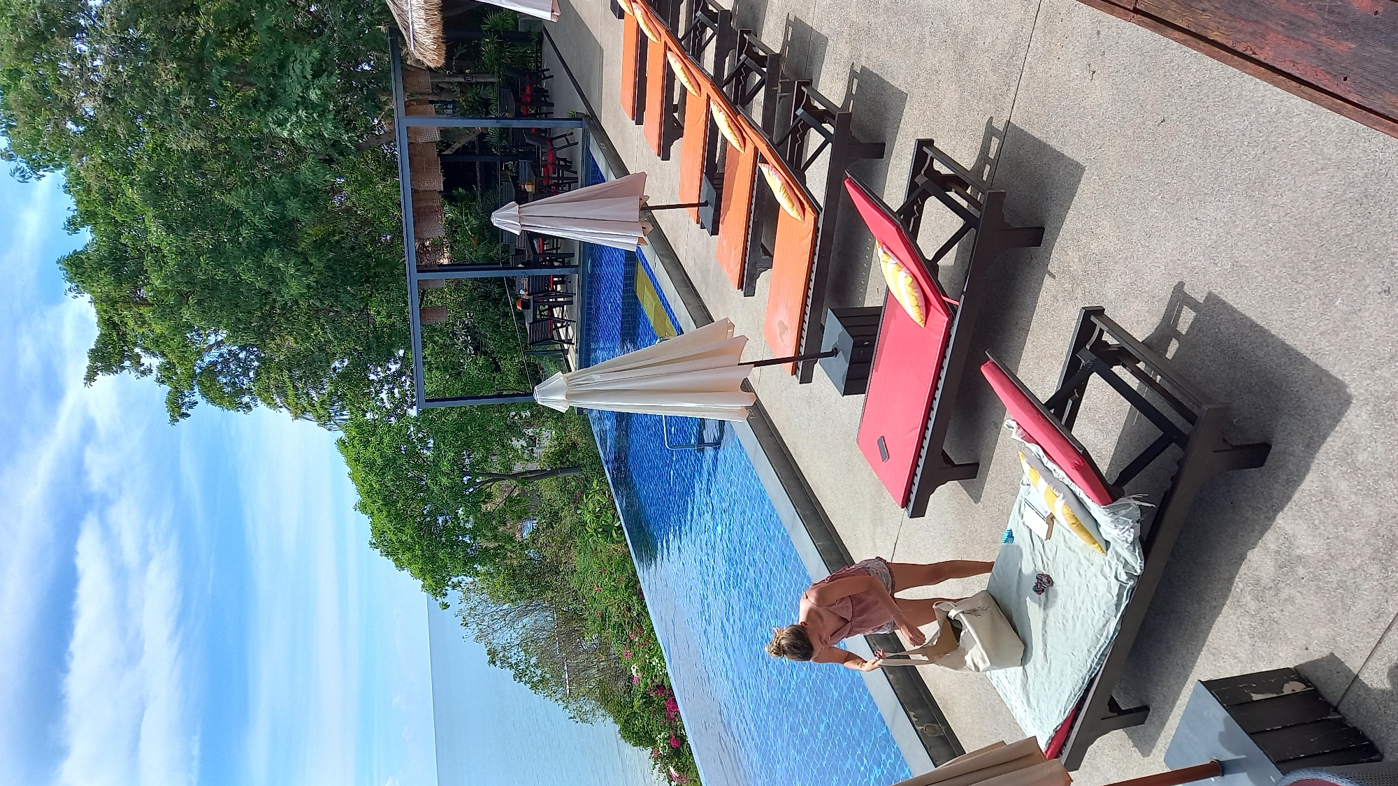 A woman with blonde hair pulled back in a bun, wearing a pink tank top and denim shorts, stands near the edge of an infinity pool in a tropical setting. She is reaching into a large, light-colored tote bag, her left hand inside. In front of her are several empty lounge chairs with colorful cushions and closed, light-colored umbrellas. The pool is filled with blue water and has a tiled bottom. Beyond the pool, lush green trees and vegetation cover a hillside, with the ocean visible in the distance under a partly cloudy blue sky. Further back, a covered seating area with tables and chairs is visible amongst the trees. The overall scene suggests a resort or hotel environment in ตำบลเกาะพะงัน, Thailand.