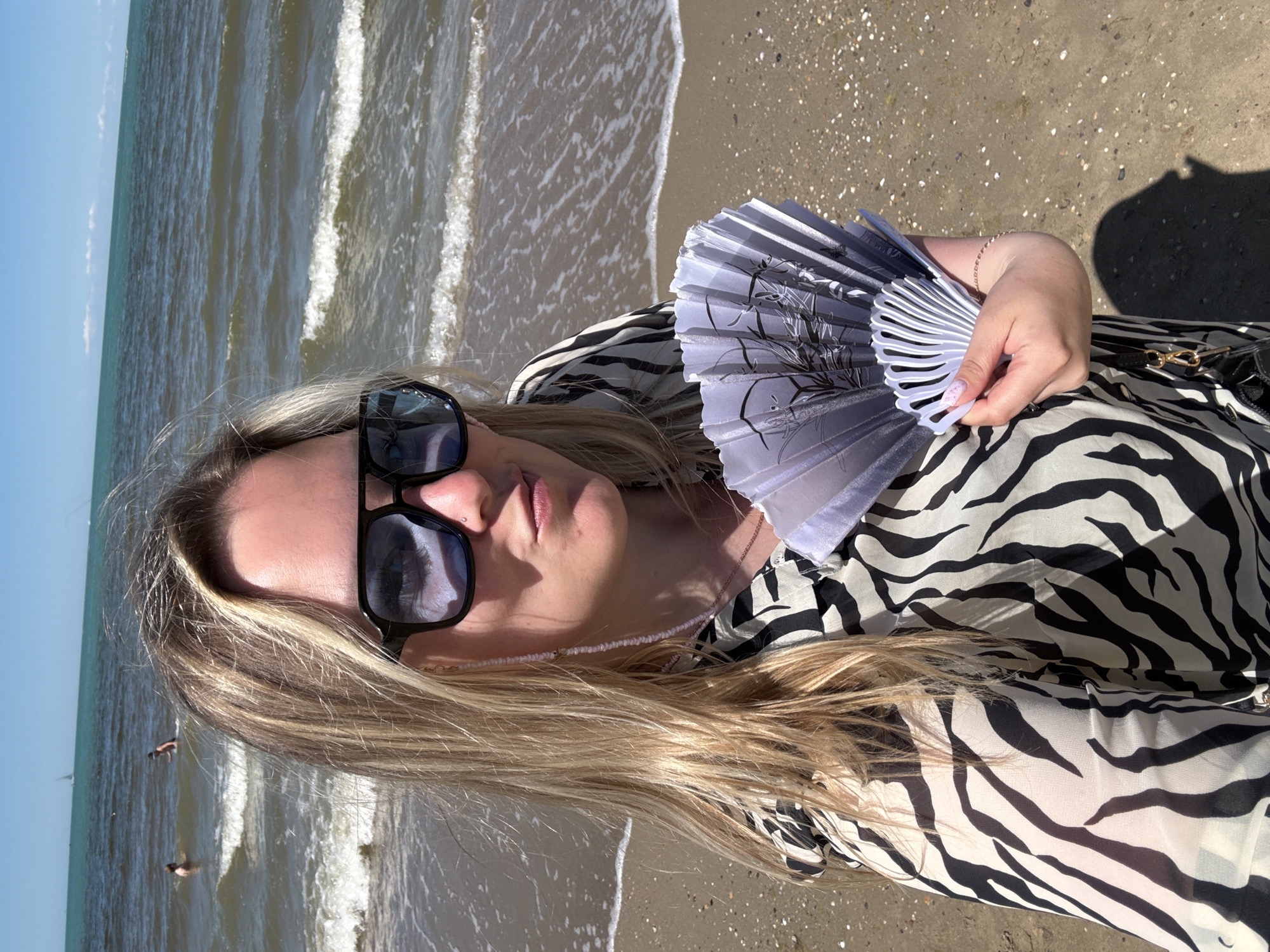 The image, rotated 90 degrees counter-clockwise, features a woman posing on a sandy beach under a clear sky in Anapa, Russia. She is positioned in the foreground, seen from the chest up, with long blonde hair. She wears dark black sunglasses that reflect the sky and distant elements. Her expression is neutral, and her lips are slightly parted.
The woman is dressed in a black and white zebra-print top or dress. Around her neck, a delicate, light pink beaded chain is visible. She holds a white, pleated hand fan adorned with a black botanical design in her left hand. A thin, possibly rose gold, bracelet encircles her left wrist, and her fingernails have a light pink polish with a subtle pattern. A black strap with a gold buckle, possibly from a bag, is visible near her hip.
The background shows the tranquil sea meeting the sandy shore. Gentle white waves break on the beach. The ocean extends to the horizon, where it meets a bright, clear blue sky with minimal cloud cover. In the distance, two small dark figures, appearing to be people, are visible in the water, along with a very faint, distant object resembling a small boat or buoy.
The scene depicts a sunny, warm day, likely late morning or early afternoon, given the bright light and distinct shadows on the sand. The overall impression is one of relaxation by the seaside. There is no visible text in the image.