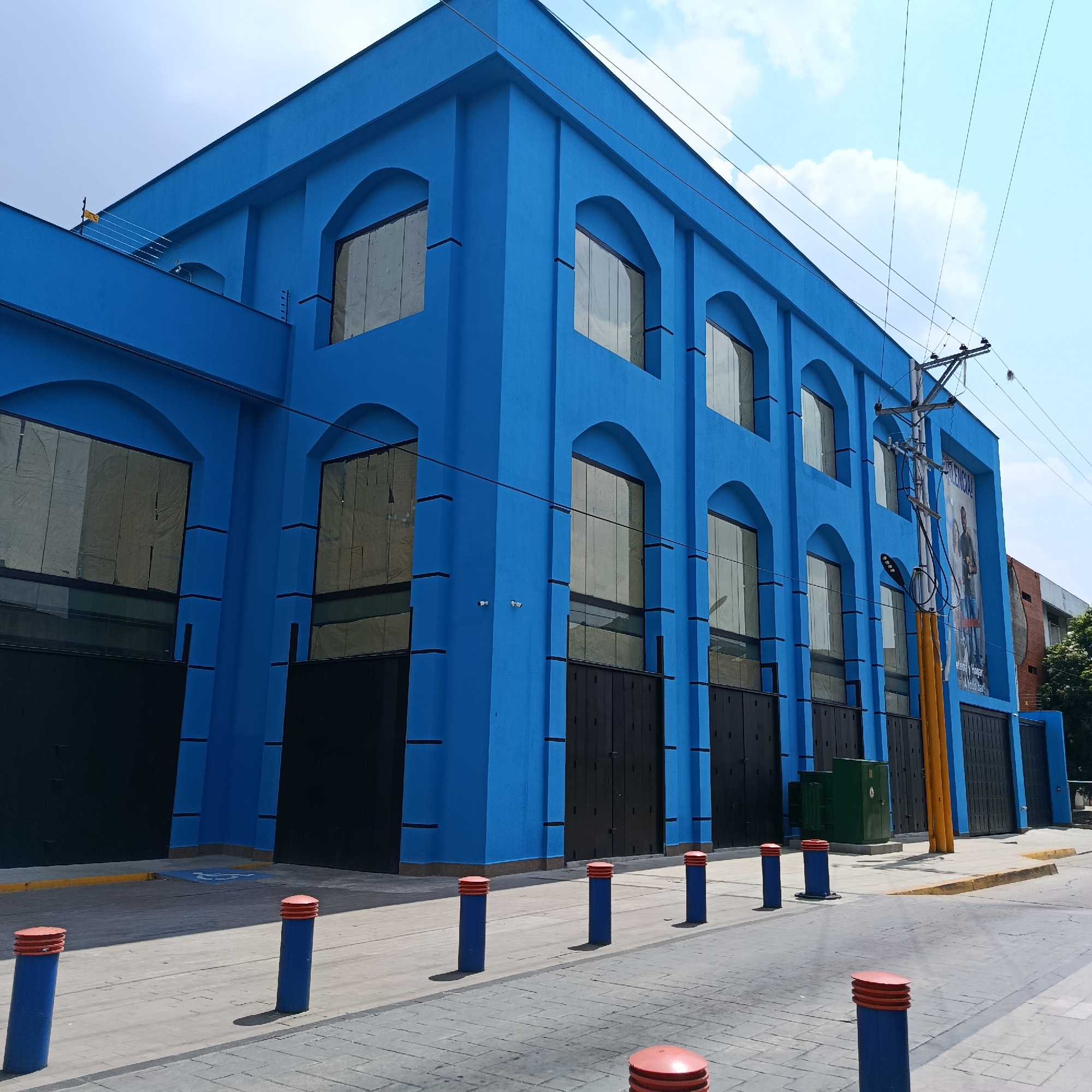 A low-angle, eye-level shot captures a large, bright blue building with a symmetrical facade. The building features two stories with arched windows on both levels. Black rectangular doors are located at the ground level beneath each window. A utility pole stands in front of the building with exposed wires running across the top of the structure. The street in front of the building is paved with rectangular stones and lined with blue cylindrical bollards with red tops. A yellow line and a handicapped parking symbol are painted on the pavement in front of the building. A green electrical box is positioned to the right of the utility pole. A poster can be seen on the right side of the building. The sky above the building is blue with scattered clouds. The location is in the city of Unknown, Unknown.