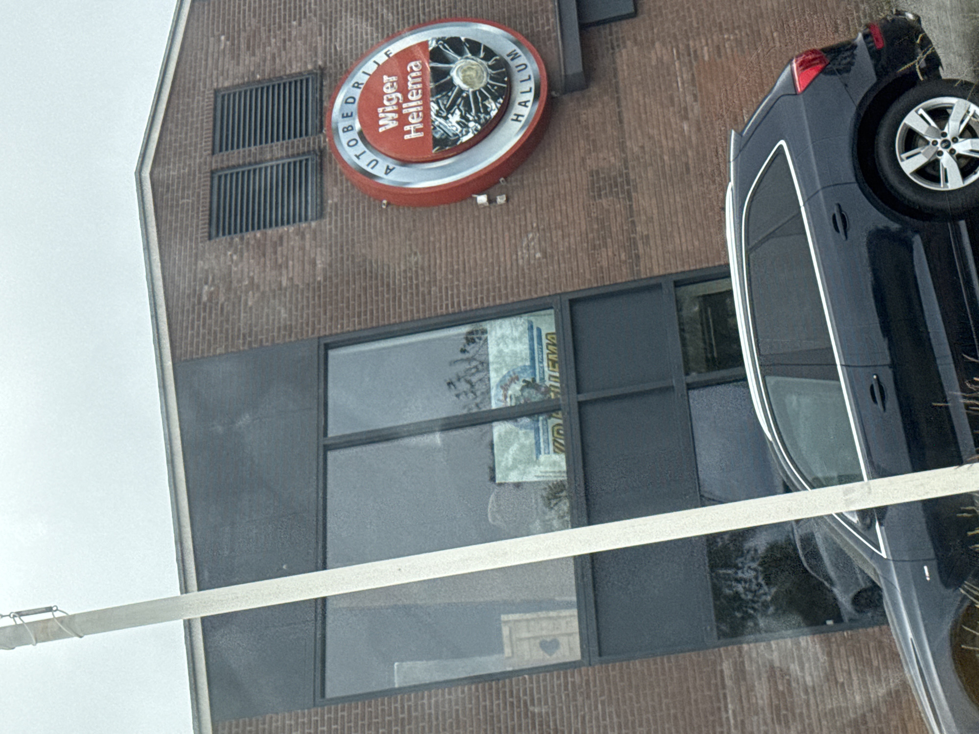 An exterior, eye-level, medium shot captures a dark-colored SUV parked in front of a brick building with large, dark-tinted windows. The building has a prominent, circular red and silver sign that reads "AUTOBEDRIJF Wiger Hellema HALLUM". The sign features an image of a car wheel. Two horizontal window vents are visible on the brick facade above the sign. A white pole, leaning at an angle, extends from the bottom left of the frame towards the top center. The sky is overcast.