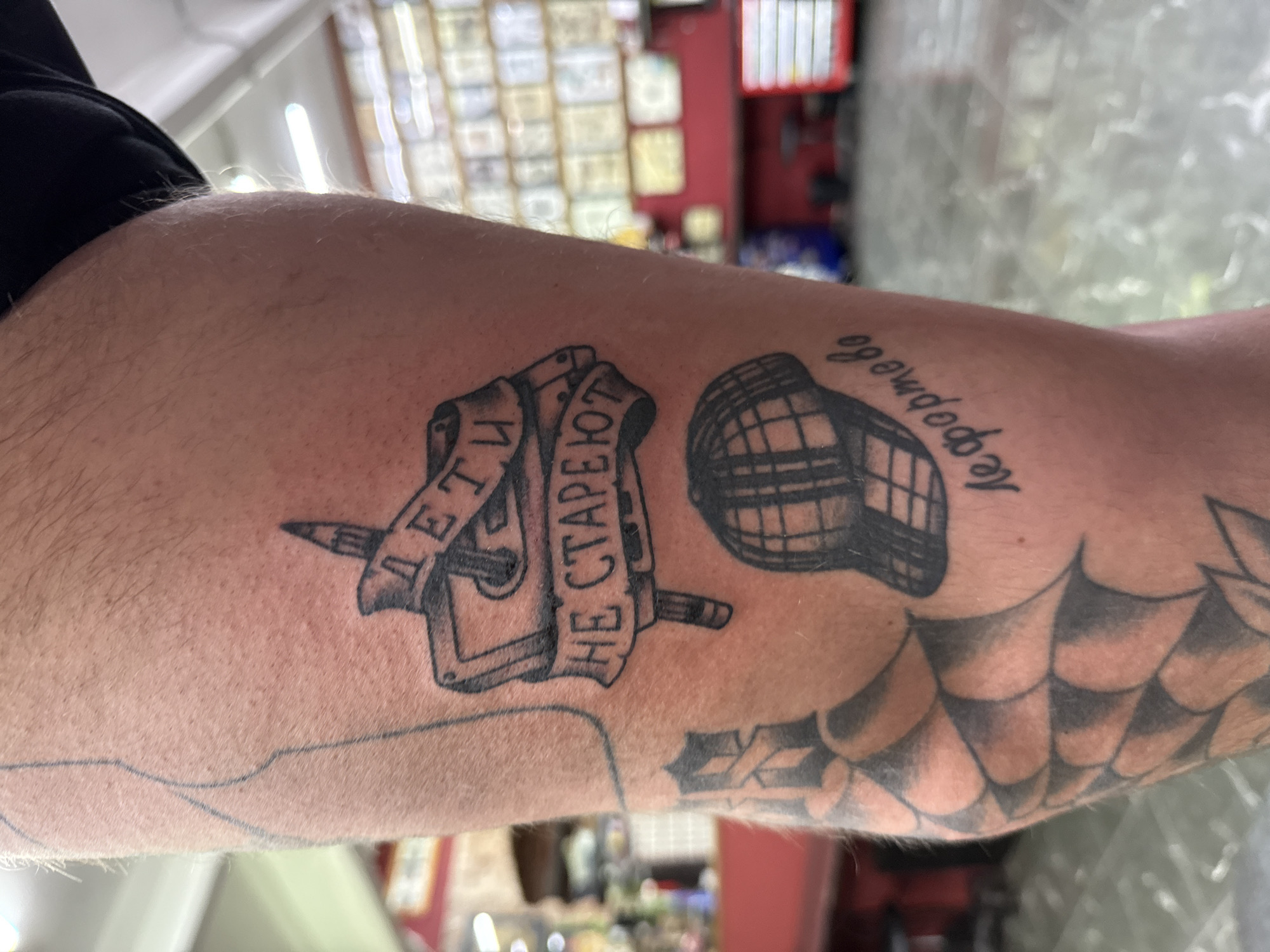 An overhead close-up shot captures a person's tattooed forearm in what appears to be a tattoo studio in Moscow, Russia. The arm, covered in light hair, displays several tattoos.
The most prominent tattoo features a banner wrapped around a stylized dagger or spike. On the banner, Cyrillic text reads "ДЕТИ НЕ СТАРЕЮТ," which translates to "Children don't grow old" or "Children do not age." To the right of this, another tattoo depicts a detailed, grid-like hockey helmet. Adjacent to the helmet, more Cyrillic script is visible, spelling "неформально," meaning "informally" or "non-formally." Further down the arm, towards the elbow, a partial tattoo with a dark, shaded scale or spiderweb pattern is visible. Below the banner tattoo, there's also a faint blue outline, suggesting a new tattoo design or a section of a larger sleeve in progress.
The background is out of focus but shows bright artificial lighting from the ceiling and shelves or display boards on the walls, typical of a tattoo parlor. Some red elements are also discernible in the blurred background. The image focuses entirely on the arm and its tattoos, with no other subjects or explicit activity. The lighting suggests an indoor setting, likely during the day or evening.