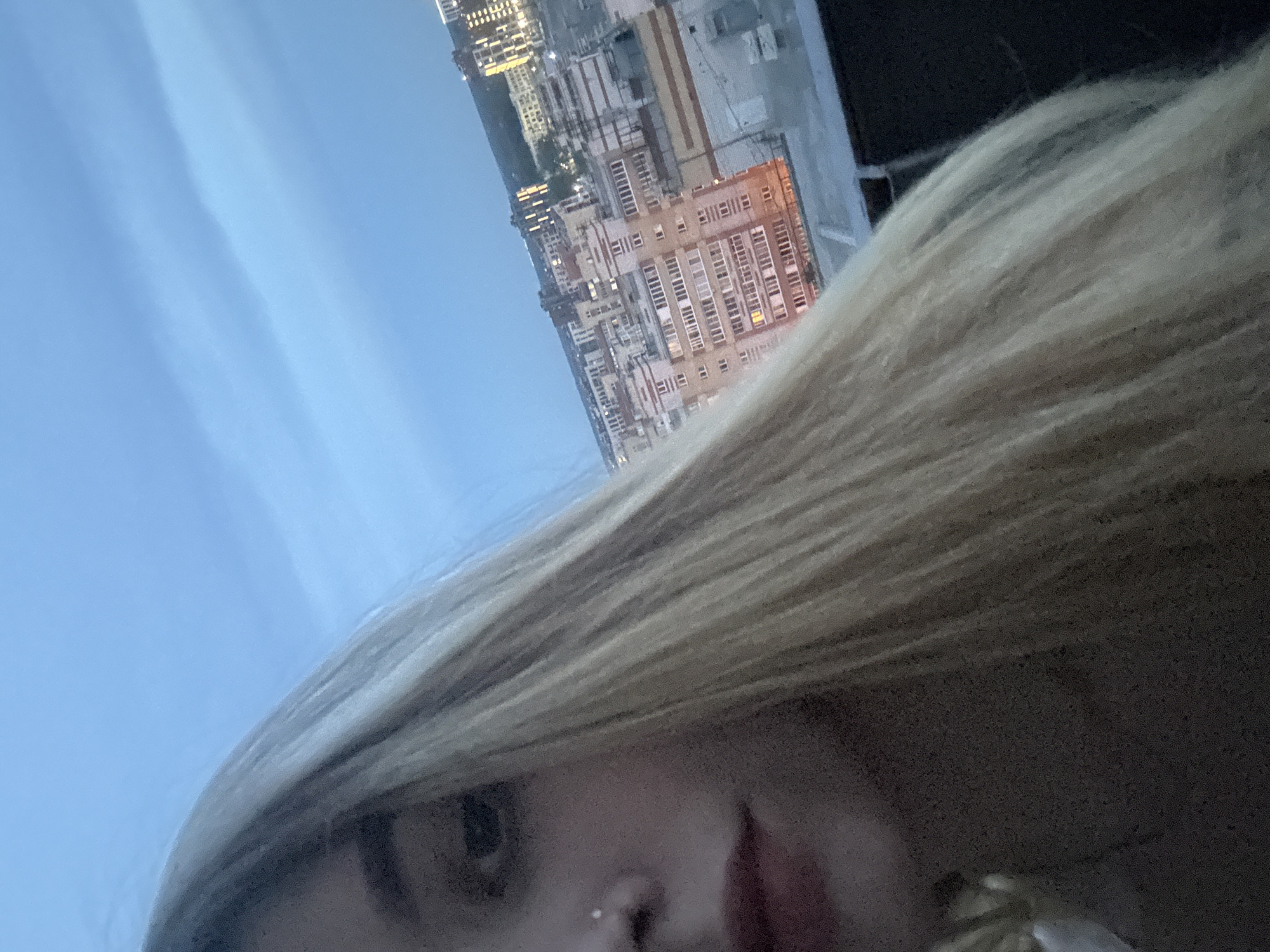 A young woman with blonde hair and a nose ring is taking a selfie outdoors at dusk. She is positioned on the left side of the frame, with her face in profile. Her eyes are closed, and her lips are slightly parted. The background features a wide expanse of the sky, transitioning from a light blue at the top to a darker blue towards the horizon, with subtle cloud formations. Below the sky, a cityscape is visible, characterized by a dense cluster of multi-story buildings with lit windows. These buildings appear to be apartment complexes, suggesting an urban environment. The overall impression is that of a residential area within a city, possibly Omsk, Russia, given the architectural style of the buildings which is common in Russian cities. The lighting indicates it is either dawn or dusk.