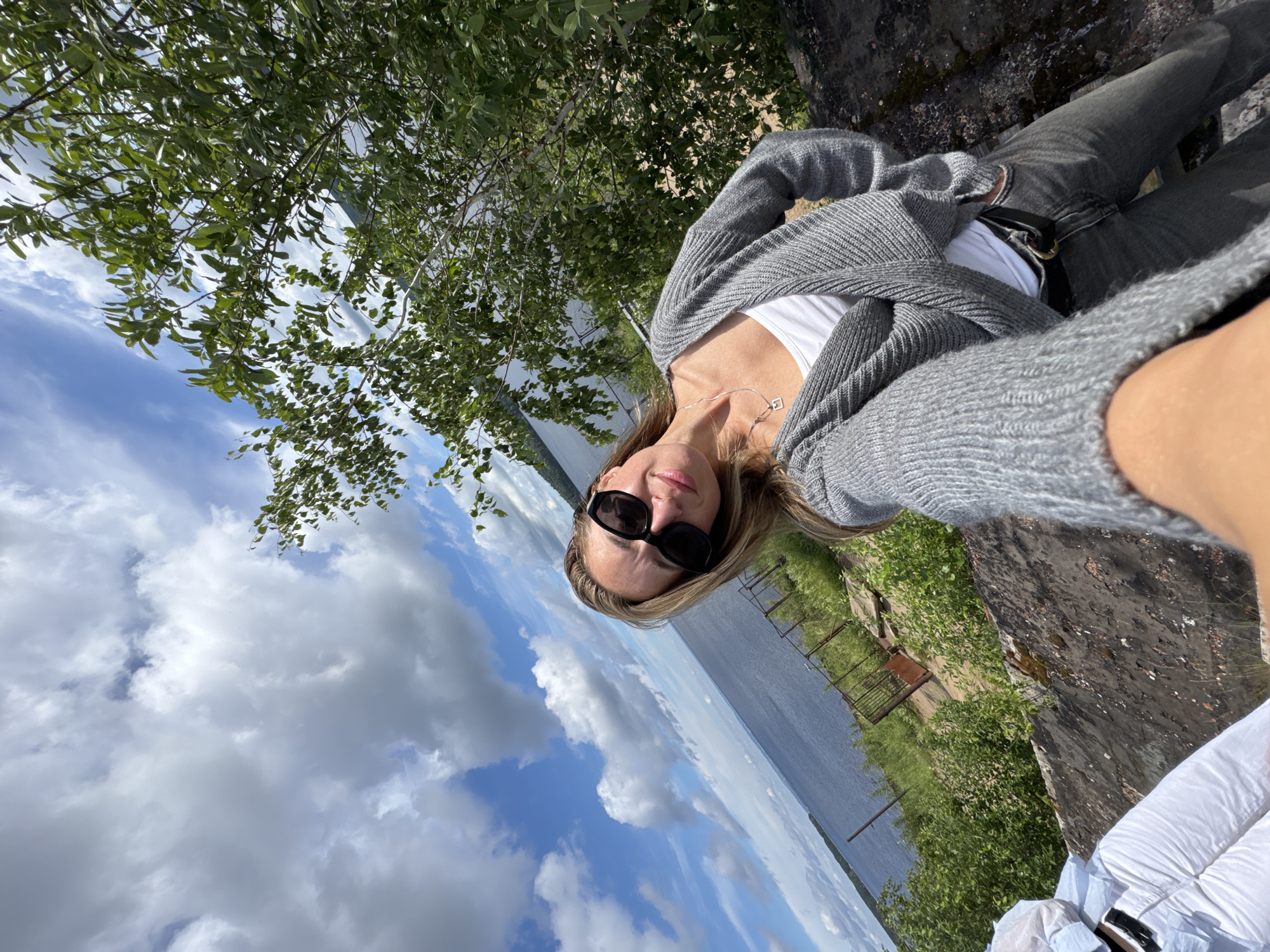 The image shows a woman taking a selfie outdoors. She is wearing a grey cardigan over a white top, grey jeans, and black sunglasses. She has blonde hair and is smiling at the camera. In the background, there is a body of water, trees, and a cloudy sky. The location appears to be a scenic spot overlooking a lake or river. Based on the presence of the Kama River and the general landscape, this could be in or near the city of Chaikovsky, Russia, a city known for its hydroelectric power station and its location on the Kama River.