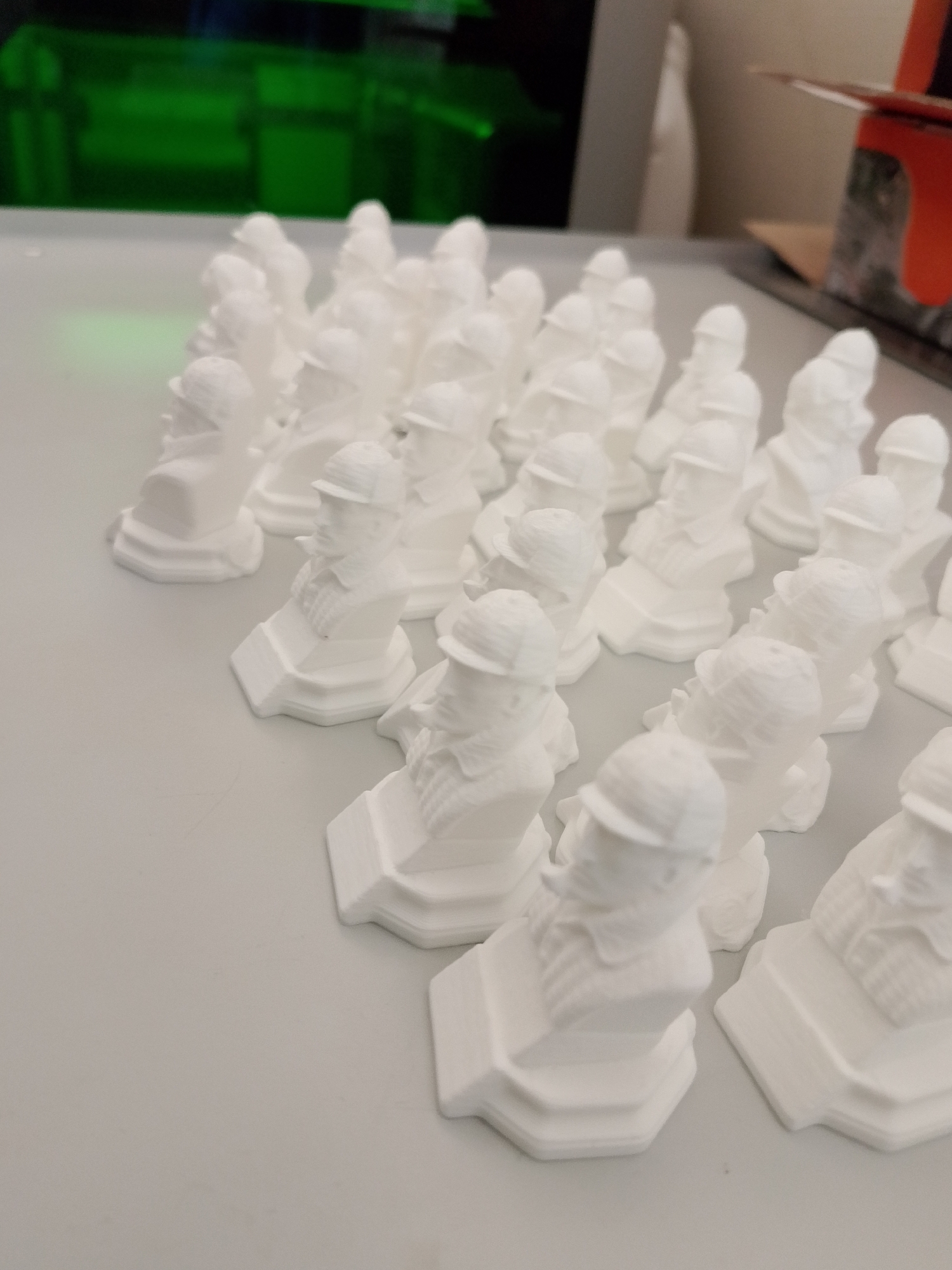 The image shows a large group of identical, small, white, 3D-printed busts arranged in rows on a light grey surface. The busts depict a man with a distinct hat and facial features. The background shows indistinct shapes and a green light source. The location in the city of Izhevsk, Russia, cannot be determined from the image alone.