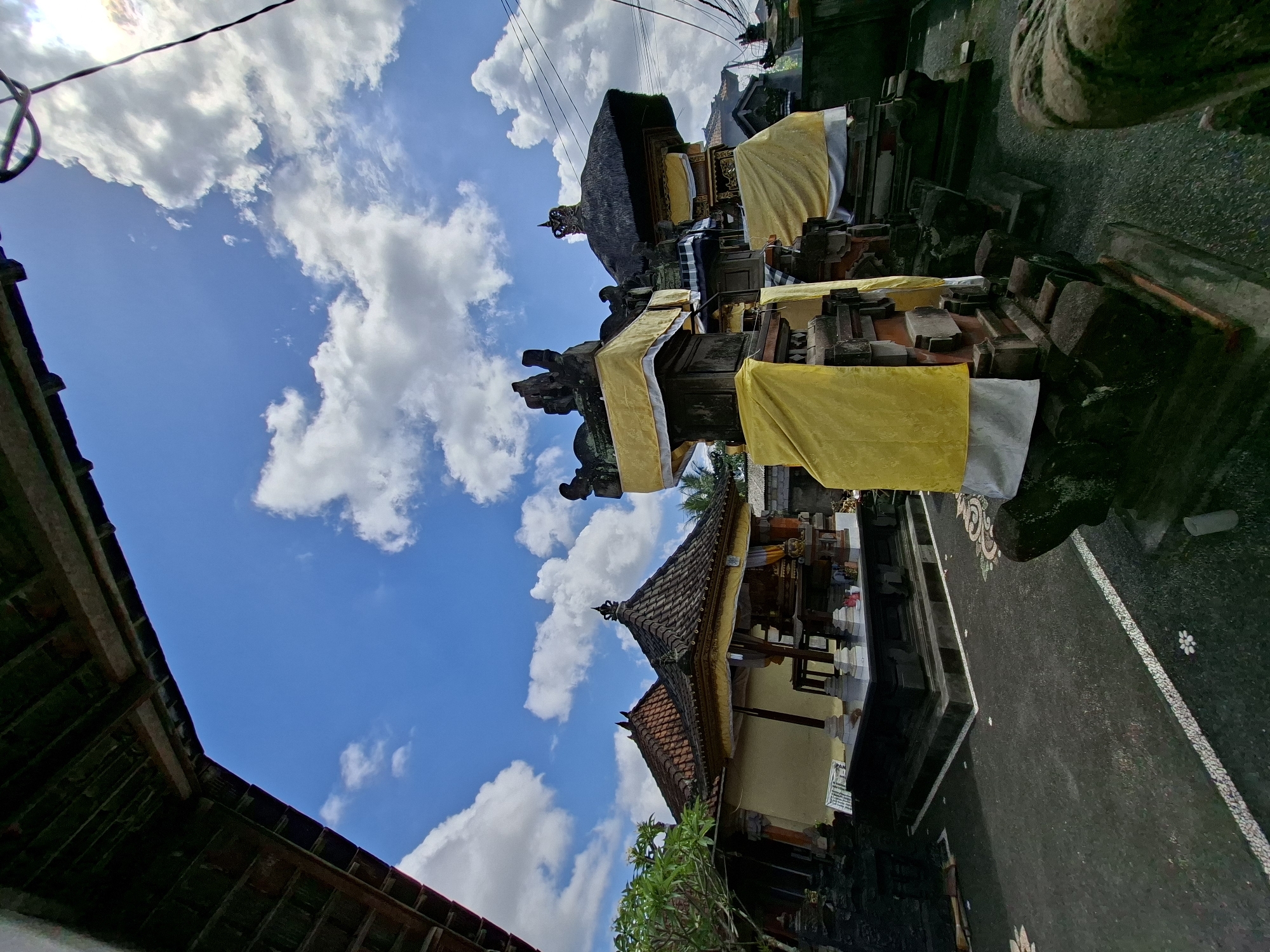 A low-angle shot captures an outdoor courtyard in Blahbatuh, Indonesia, featuring traditional Balinese architecture under a bright blue sky with scattered white clouds. In the foreground, intricately carved stone structures are draped with yellow and white fabric, suggesting a ceremonial setting. The structures are layered with stone and wood, topped with ornamental elements and statues. To the left, a building with a tiered tiled roof and yellow trim is visible, with a small tree in front. The courtyard floor is dark asphalt with decorative white borders and small floral motifs. Overhead, a portion of a wooden overhang and electrical wires are present, indicating a populated area. The scene is static and appears to be a daytime photograph.