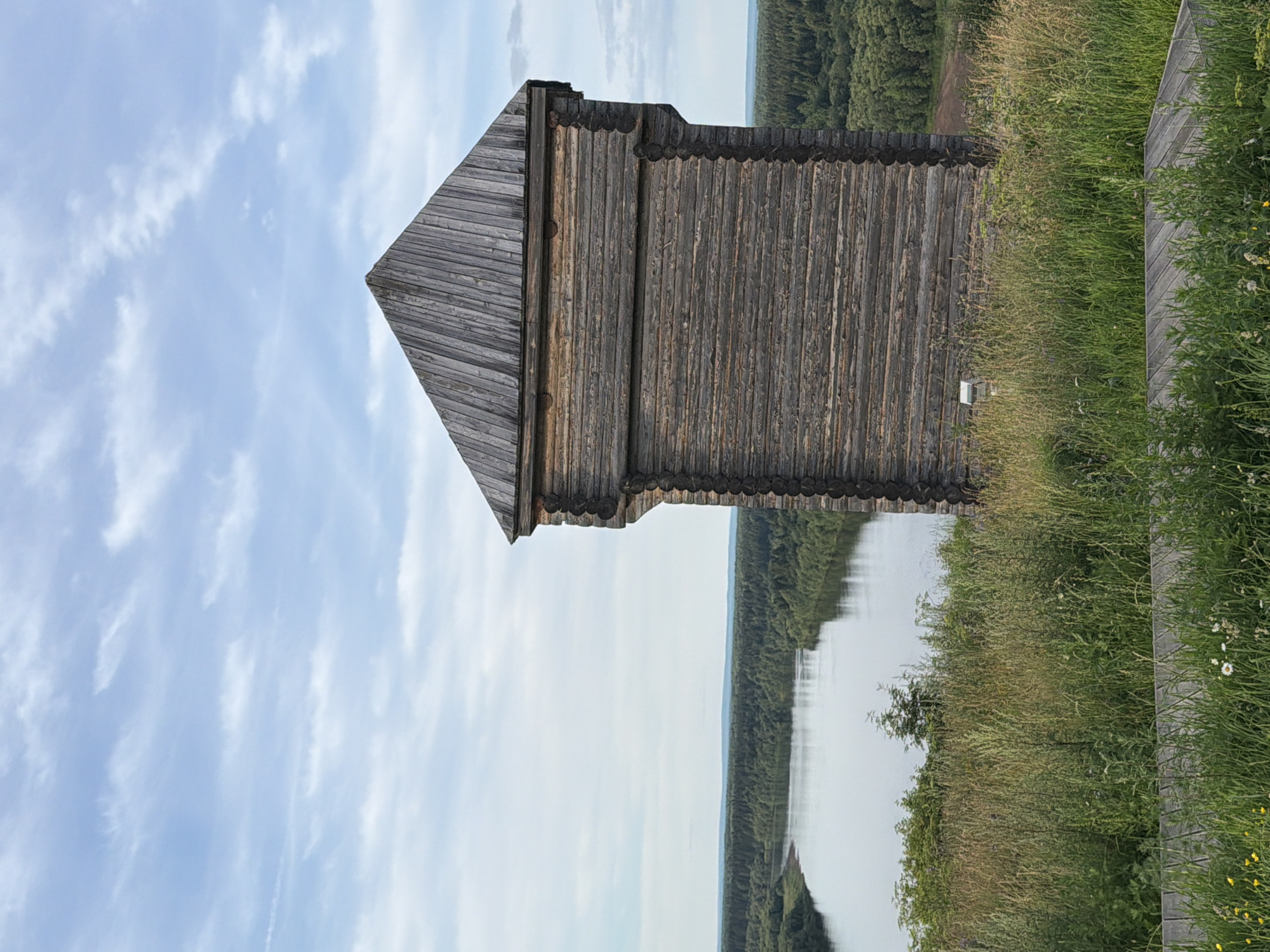 The image is a vertical, medium shot of a traditional wooden tower structure. The tower, made of weathered grey logs, has a pointed, roof-like section and intricate decorative detailing along its upper edge. To the left of the tower, the sky is a pale blue with scattered white clouds. To the right of the tower, a body of calm water reflects the distant treeline, which appears as a dense, dark green forest. The foreground is dominated by tall, wild grasses and wildflowers, with a wooden walkway or railing visible on the far right. The scene suggests a natural, outdoor setting, likely a historical or scenic location. The lighting indicates it is daytime, with soft, diffused sunlight. No people are visible, and there is no discernible text in the image. The overall impression is one of rustic architecture set against a serene natural landscape.