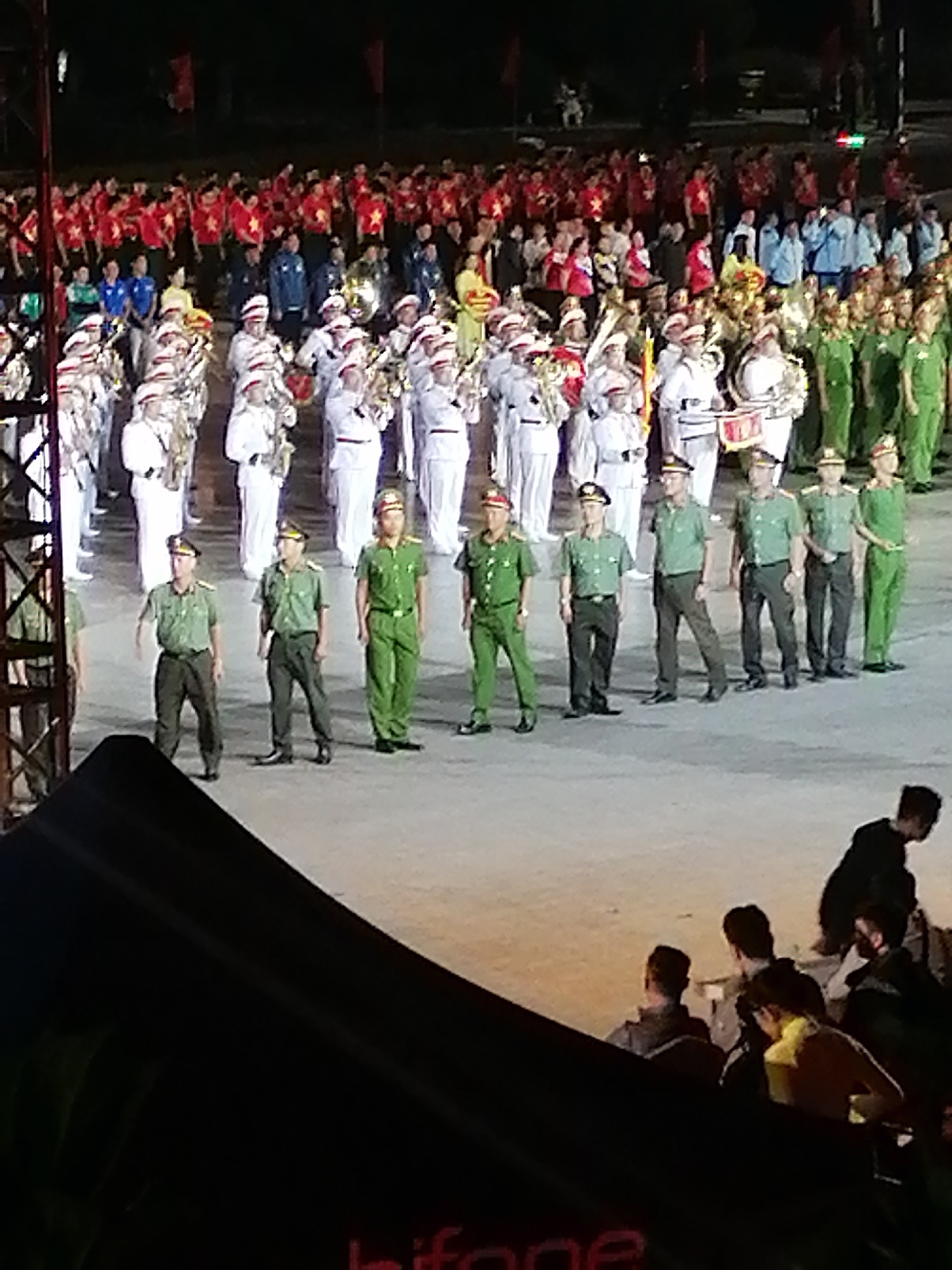 The image depicts a large group of people, many of whom are dressed in military-style uniforms, possibly participating in a parade or ceremony. The setting appears to be an outdoor event, with a dark background suggesting nighttime or a covered venue. Several individuals in the foreground are wearing green uniforms and caps, standing in formation. Behind them, a band or orchestra is visible, with some members holding instruments. Further in the background, a crowd of onlookers, many in red shirts, are gathered. The presence of a stage-like structure or platform is also suggested by the foreground elements. The image quality is somewhat degraded, with a grainy appearance. The context of Da Lat, Vietnam, suggests this could be related to a national holiday or significant event.