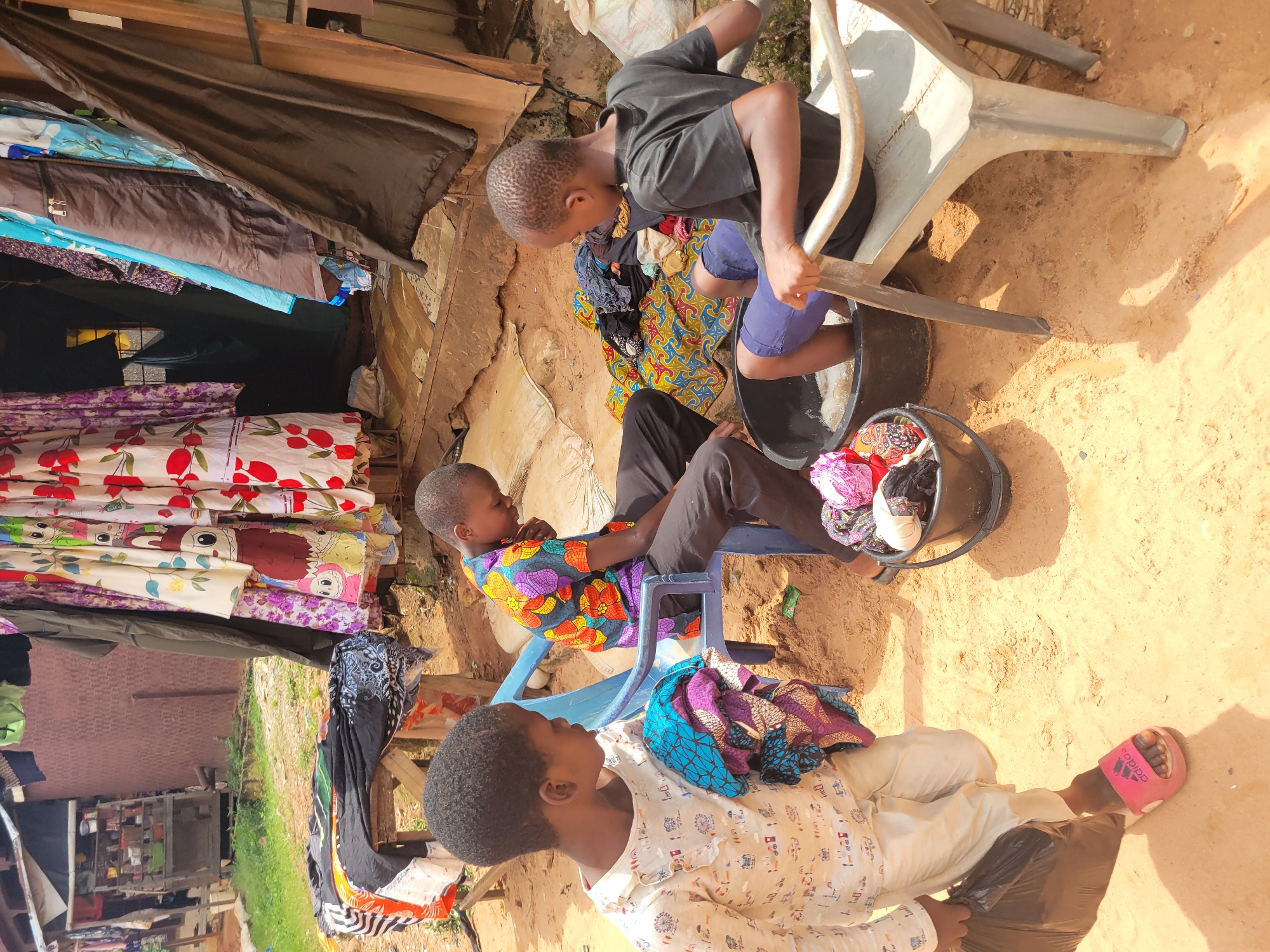 Three young individuals, likely children, are engaged in laundry activities in an outdoor, sandy environment, possibly a market or residential area in Onitsha, Nigeria. In the foreground to the left, a child wearing a white long-sleeved shirt with a graphic print and light-colored pants is holding a black plastic bag and a bundle of colorful fabrics. To the right, another child in a vibrant, patterned short-sleeved shirt and dark pants sits on a blue plastic chair, looking down. In the center right, a third child, wearing a dark t-shirt and blue shorts, is seated on a light-colored plastic chair, with their feet immersed in a large black basin filled with soapy water, indicating they are washing clothes. Scattered around the basin are more colorful fabrics and a smaller black bucket containing laundry. The background features a rustic stall with hanging garments, shelves stocked with goods, and wooden structures, suggesting a marketplace setting. The ground is primarily sand, with some areas of dry dirt and sparse vegetation. The lighting indicates it is daytime, likely under direct sunlight, casting shadows.