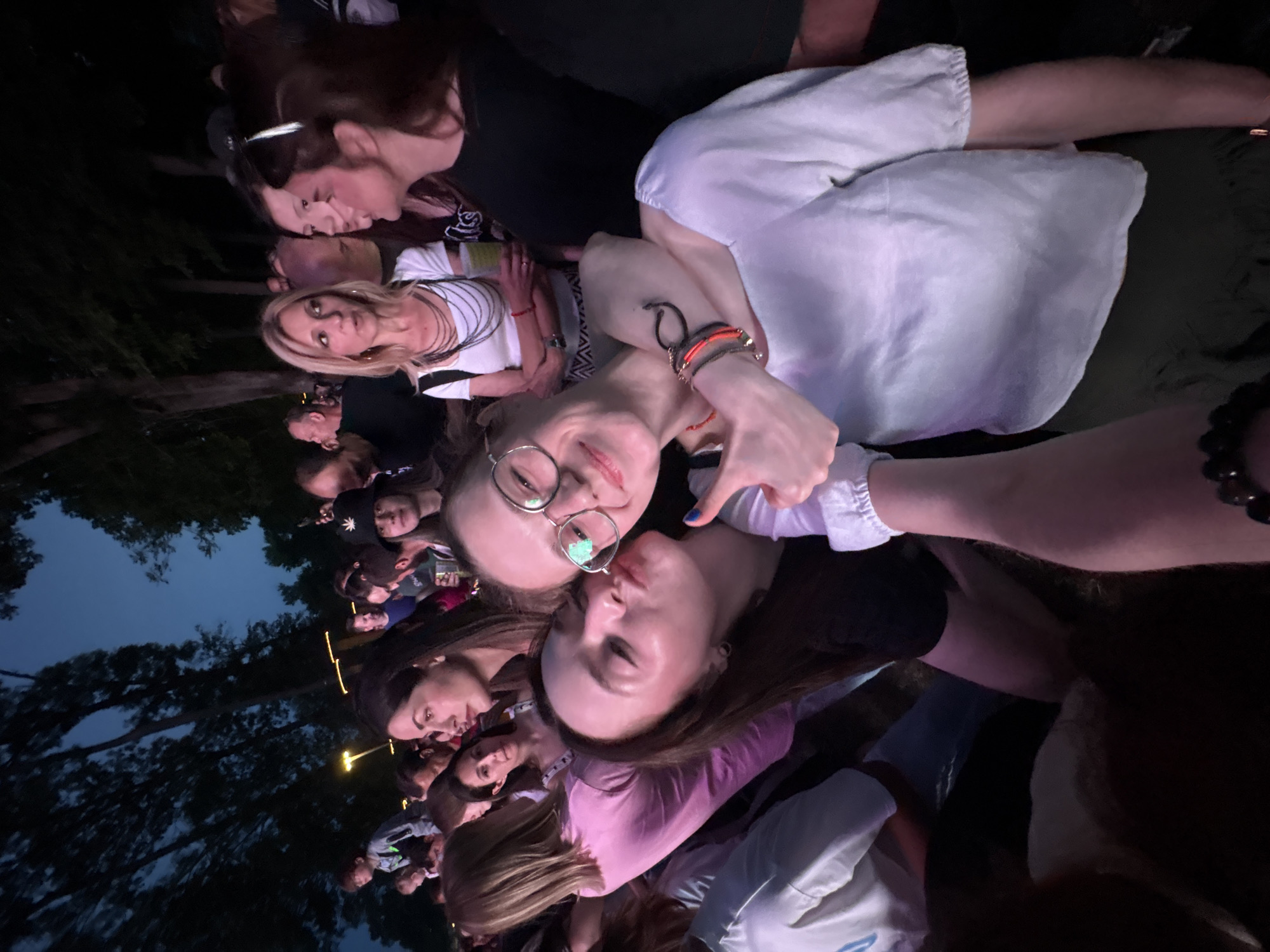 The image shows a crowd of people, seemingly at an outdoor event at night. The location appears to be in Kragujevac, Serbia. Two young women in the foreground are posing for the camera, one wearing glasses and the other with a pink shirt. The woman in glasses is making a playful gesture. The background consists of other attendees, some of whom are also looking at the camera. The lighting suggests an evening setting with artificial lights. The atmosphere seems lively.