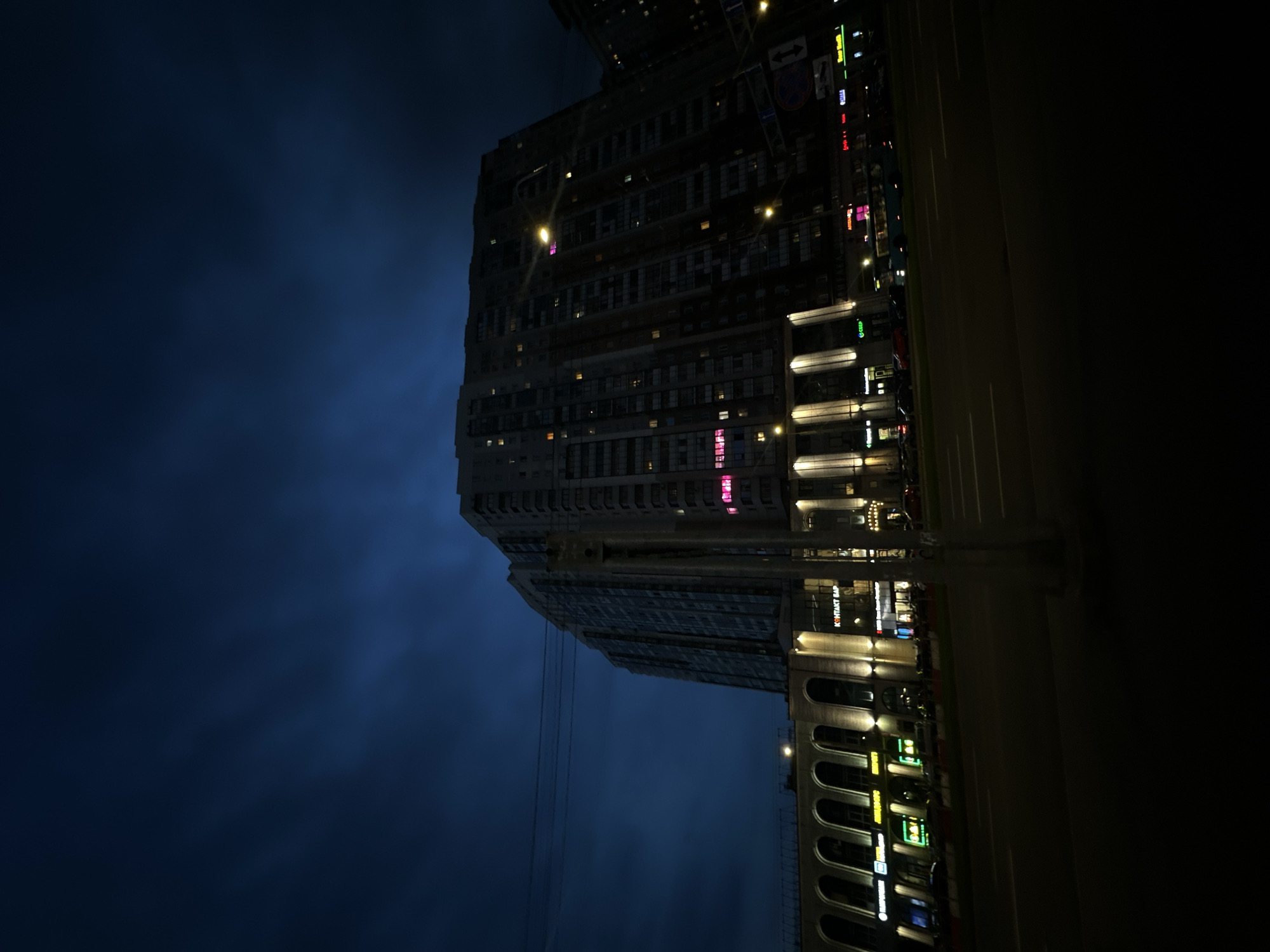 The image captures an urban street scene at night. The sky is a dark, deep blue with visible cloud cover. A tall, multi-story building dominates the background on the right, featuring numerous rectangular windows. Many of these windows exhibit internal yellow-orange illumination, while several others display a distinct pink or magenta light. Below and to the left of this tall structure, a lower commercial building extends horizontally. This building incorporates a series of regularly spaced arched openings on its upper level, each highlighted by a warm-toned upward-facing spotlight.
The ground floor of the commercial building houses multiple businesses, indicated by illuminated signs. Visible Cyrillic text on these signs includes "КОНТАКТ БАР" (Kontakt Bar), "АПТЕКА" (Pharmacy) on a pink sign, and "СБЕР" (Sber) on a green sign. Other storefront signs are present with various colors of illumination (green, yellow, white), but their text is not clearly legible.
In the foreground, a multi-lane asphalt road is visible with white painted lane markings, including solid and dashed lines. A dark utility pole stands vertically in the mid-foreground, slightly to the left of center, partially obscuring the view of the buildings behind it. Overhead utility wires stretch horizontally across the upper-left portion of the sky. Multiple streetlights, emitting bright yellow-orange light, illuminate the street and parts of the buildings. Road signs on poles are present on the right side of the street, but their details are indistinct.
Several vehicles are observed: a line of parked cars is situated along the curb in front of the commercial establishments, and a light-blue public bus is seen in the far lane, traveling away from the camera. No individuals are visible anywhere in the scene. The location is an unknown city.