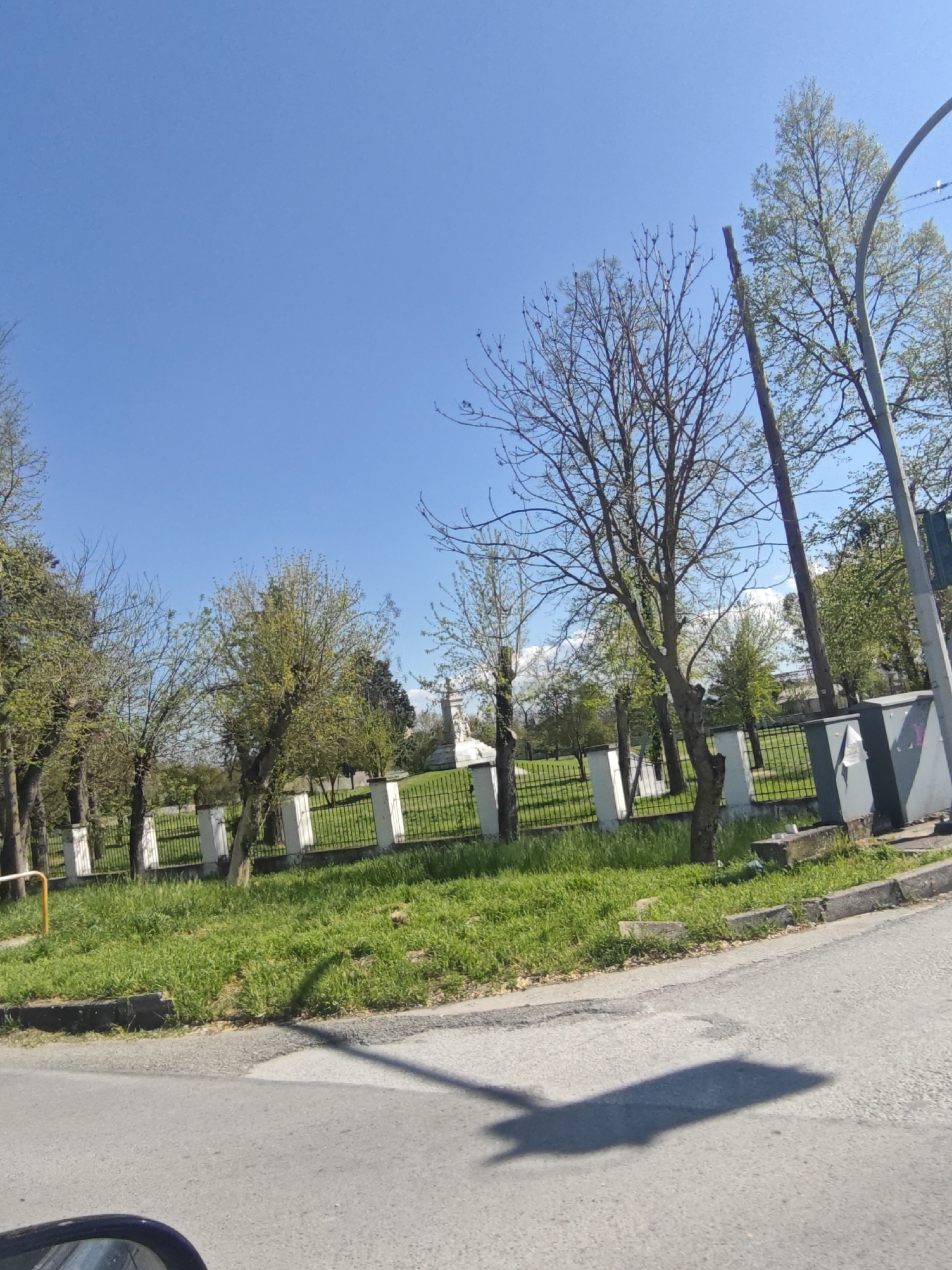 The image shows a street scene in Serres, Greece. A paved road curves to the right of the frame, with part of a vehicle visible in the lower left corner. To the left of the road is a grassy area enclosed by a low black metal fence with white pillars. Trees with sparse foliage are scattered across the grassy area. In the background, a white monument is visible through the fence. Utility poles and metal boxes are situated near the fence. The sky is clear and blue. Shadows are cast on the road surface.
