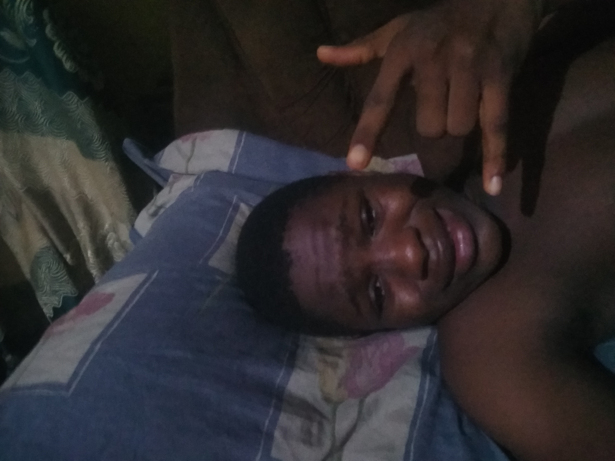 A person is lying on a bed with a patterned blue and white pillow. The person's head is resting on the pillow, and their right shoulder and part of their chest are visible. Another hand, presumably belonging to someone else, is raised above the person's head, making a hand gesture. The background is dark and appears to be a room interior. The location is Shangisha, Nigeria.