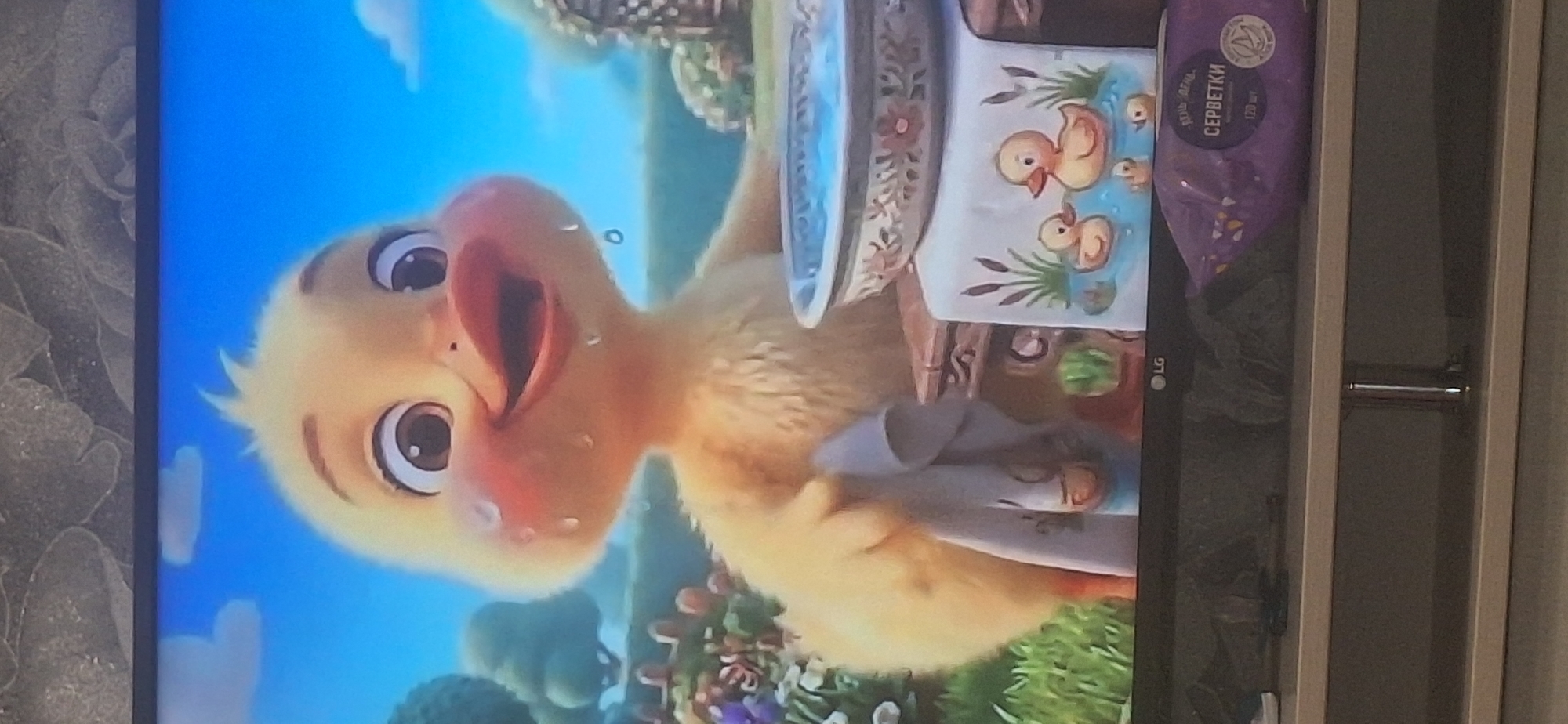 A cartoon baby duckling, with large eyes and rosy cheeks, is shown in a close-up on a television screen. The duckling has water droplets on its face and is holding a white cloth. In front of the duckling is a bowl filled with blue water and a small towel with an illustration of ducklings in water. The background shows a green grassy area with trees and flowers, indicative of an outdoor setting. The television is placed on a stand, with a purple package of wet wipes visible on the stand in front of the screen. The scene is consistent with a children's animated program. The image was taken in Bila Tserkva, Ukraine.