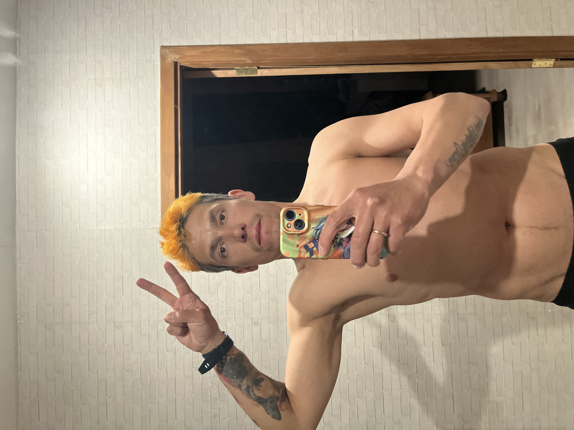 A shirtless man with orange-dyed hair is taking a selfie indoors. He is holding a phone with a patterned case and making a peace sign with his left hand. He has tattoos on his left forearm and right bicep. The background includes a white brick-patterned wall and a dark doorway framed with brown wood. There is no information in the image to determine the location in the city of Chaikovsky, Russia.