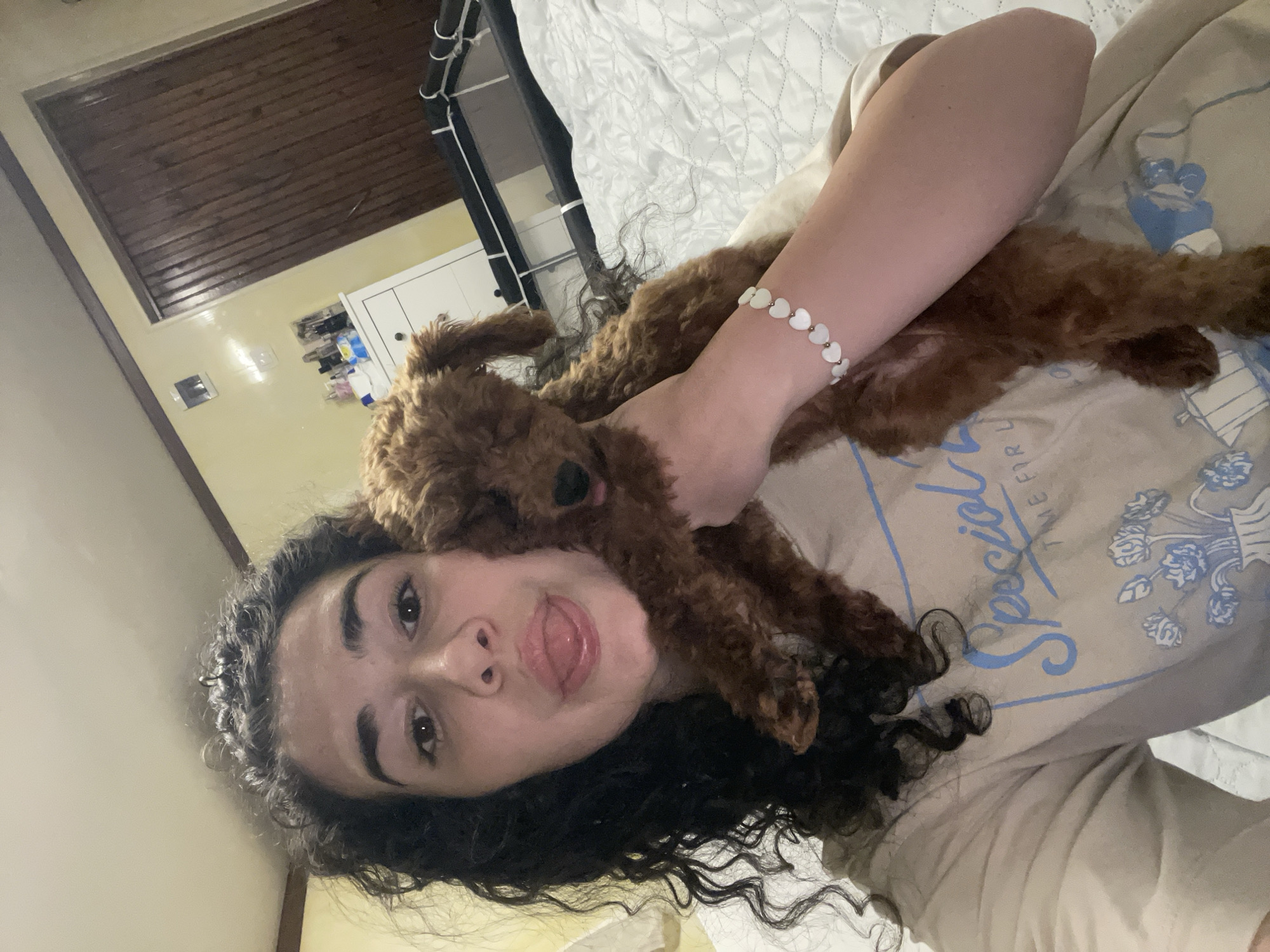 A woman is lying down with a small, brown, curly-haired dog lying on her chest. The woman is looking directly at the camera, sticking her tongue out. She has dark, curly hair and a light complexion. She is wearing a light-colored t-shirt with a blue design. She has a white bracelet with heart-shaped beads on her wrist. The background includes a bed with a white comforter, a white dresser, and a wooden ceiling. The location is a bedroom, but the city of Sofia, Bulgaria, is not identifiable from the image.