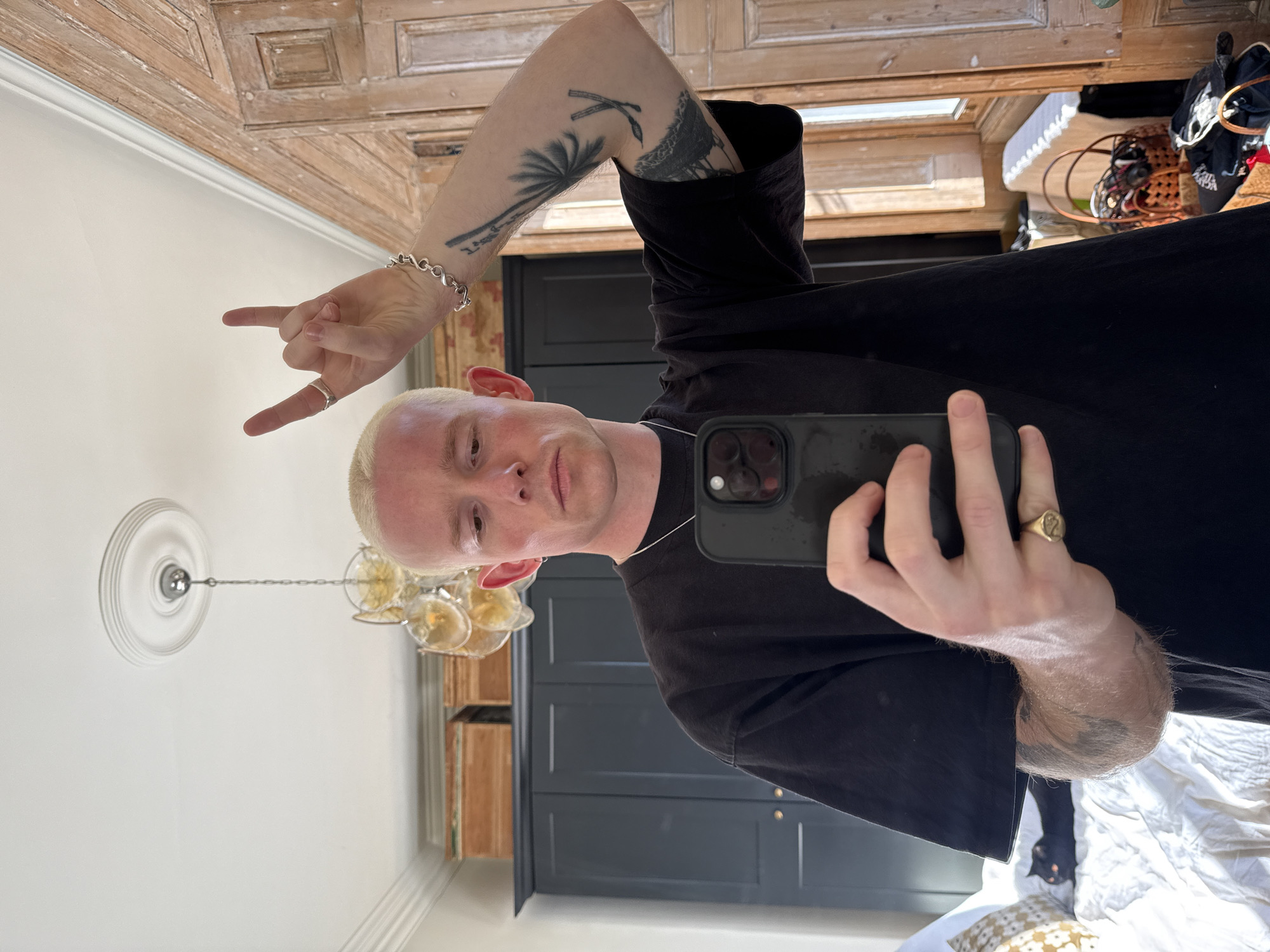 A person with short, bleached blond hair is taking a selfie in a room. The individual is wearing a black t-shirt and has multiple tattoos on their arms and hands, including a palm tree and abstract designs. They are holding a black smartphone and making the "rock on" hand gesture. The room features a wooden ceiling with a decorative pattern, a white ceiling with a chandelier, and built-in dark-colored cabinets. A bed with white bedding and patterned pillows is visible in the lower part of the frame. There are no indications that the scene is in the city of London, United Kingdom.