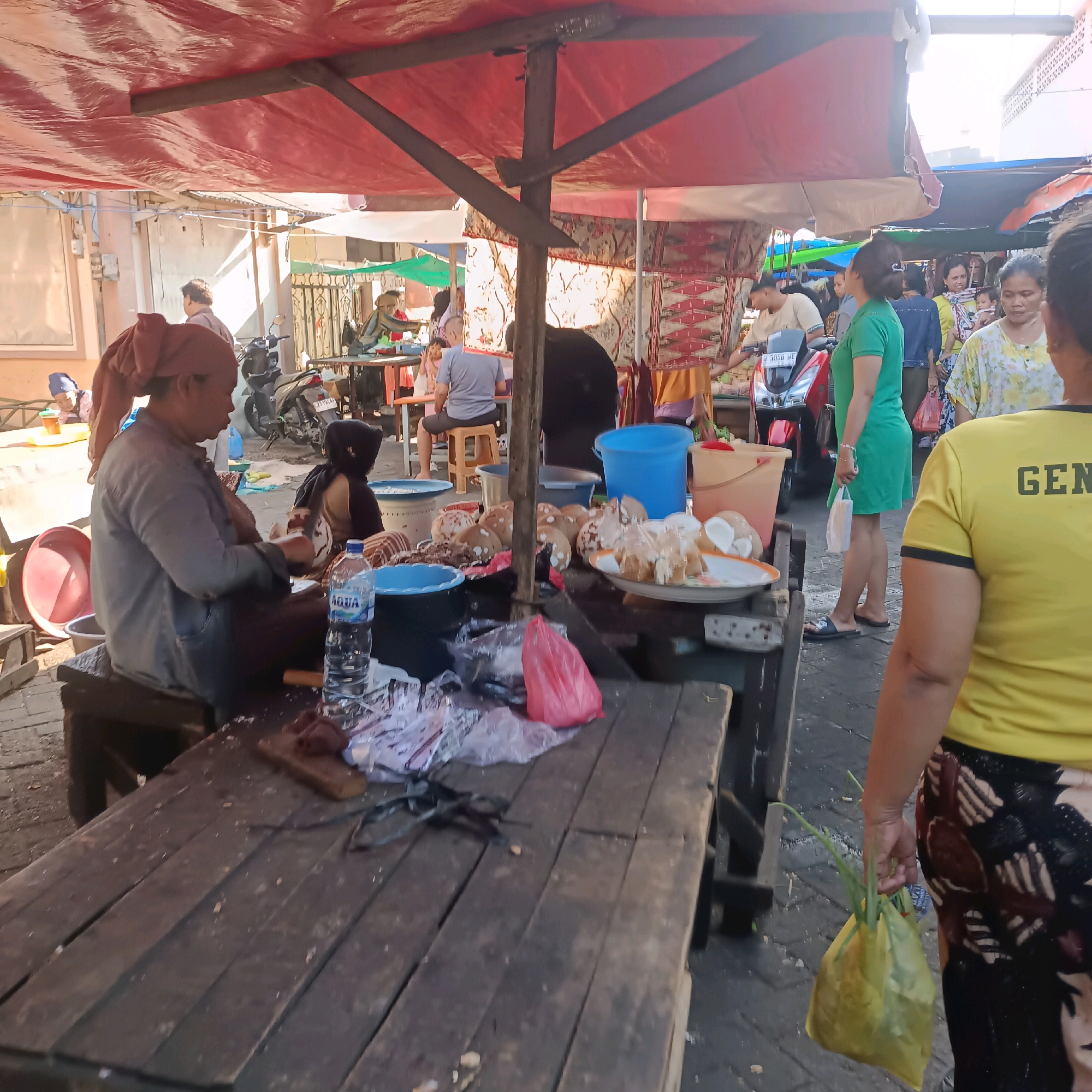 This is a medium shot of a busy outdoor market scene in Surabaya, Indonesia, likely near a street market given the presence of motorcycles and food stalls. In the foreground, a woman is seated on a low wooden stool to the left, wearing a grey long-sleeved shirt and a brown headscarf. She is focused on a wooden cutting board in front of her, where she appears to be preparing food. Next to her is a clear plastic bottle of "AQUA" brand water. A blue plastic basin and a red plastic bowl are also visible on the wooden table in front of her. In the middle ground, several coconuts are displayed on a plate, some of which have been cut open, revealing the white flesh. Plastic bags, including a pink one, are scattered around. Behind her, another person, partially visible and wearing a dark head covering, is also seated at a stall. A red motorcycle is parked in the background. The market is covered by red tarps held up by wooden poles, creating a shaded area. Other people can be seen walking and browsing among the stalls, including a woman in a bright green dress and a woman in a yellow shirt with black lettering on the back. The ground is paved with grey bricks. The overall impression is of a vibrant and bustling local market.