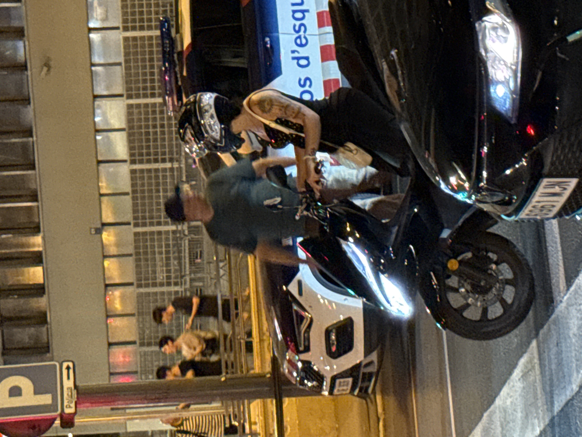 At night, on a city street in Barcelona, Spain, several people and vehicles are visible. A police vehicle, possibly a patrol car, is parked on the right side of the image. A woman wearing a helmet and a black dress is seated on a black scooter in the foreground, facing left. She has tattoos on her left arm and a small white bag slung over her shoulder. To her left, a man in a green shirt and dark baseball cap stands near another scooter. In the background, a group of people are gathered near a building with a sign that reads "Policia." Another car, white and black, is parked between the scooters and the police vehicle. The street is paved, and a white line marking a crosswalk is visible in the lower left corner. The lighting suggests an urban environment with artificial streetlights and building illumination.