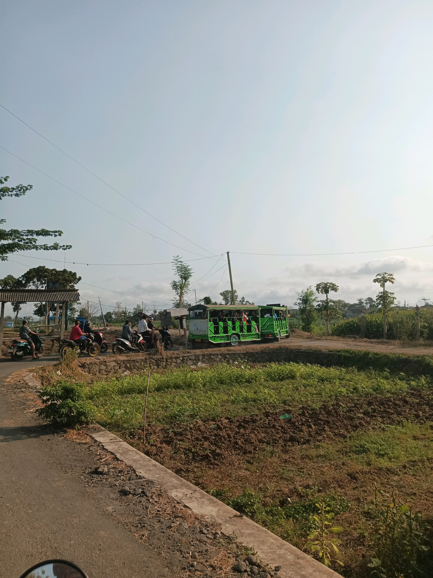 This is a vertical, medium-shot, outdoor photograph taken on a sunny day in Baruharjo, Indonesia. The scene depicts a rural road with several individuals on motorcycles and a bright green open-air vehicle.
On the left side of the road, a group of men are gathered around parked motorcycles. One man in a grey t-shirt is riding a dark-colored motorcycle, heading away from the viewer. Another man in a red t-shirt is on a red motorcycle, also facing away. Behind them, several other motorcycles are visible, some with riders. A weathered, open-sided structure with a corrugated metal roof stands to the left of the road, providing shade.
Further down the road, a vibrant green, open-sided vehicle, resembling a safari bus or a shuttle, is in motion. It appears to be carrying passengers, some of whom are visible through the open sides. The vehicle has a banner on its side with Indonesian writing.
The road itself is paved but shows signs of wear and tear, with rough patches and loose stones. It curves to the right, leading into the distance. To the right of the road, there is a field with tilled brown earth and patches of green vegetation. Beyond the field, more greenery, trees, and rural structures are visible under a clear, light blue sky with scattered white clouds. Power lines are strung across the sky. The overall impression is of a typical day in a rural Indonesian setting.